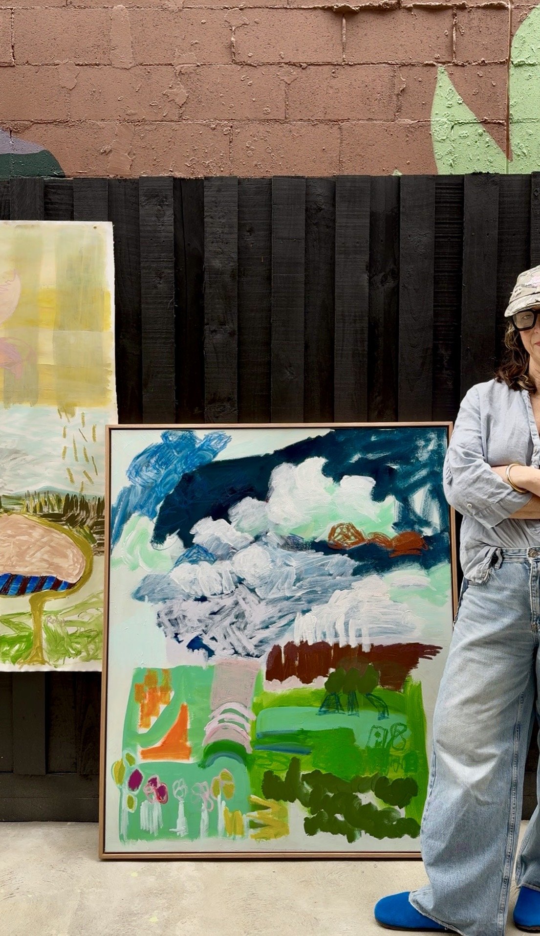 Colorful abstract paintings displayed outdoors with a brick and wooden fence background, partially showing a woman in casual clothing.