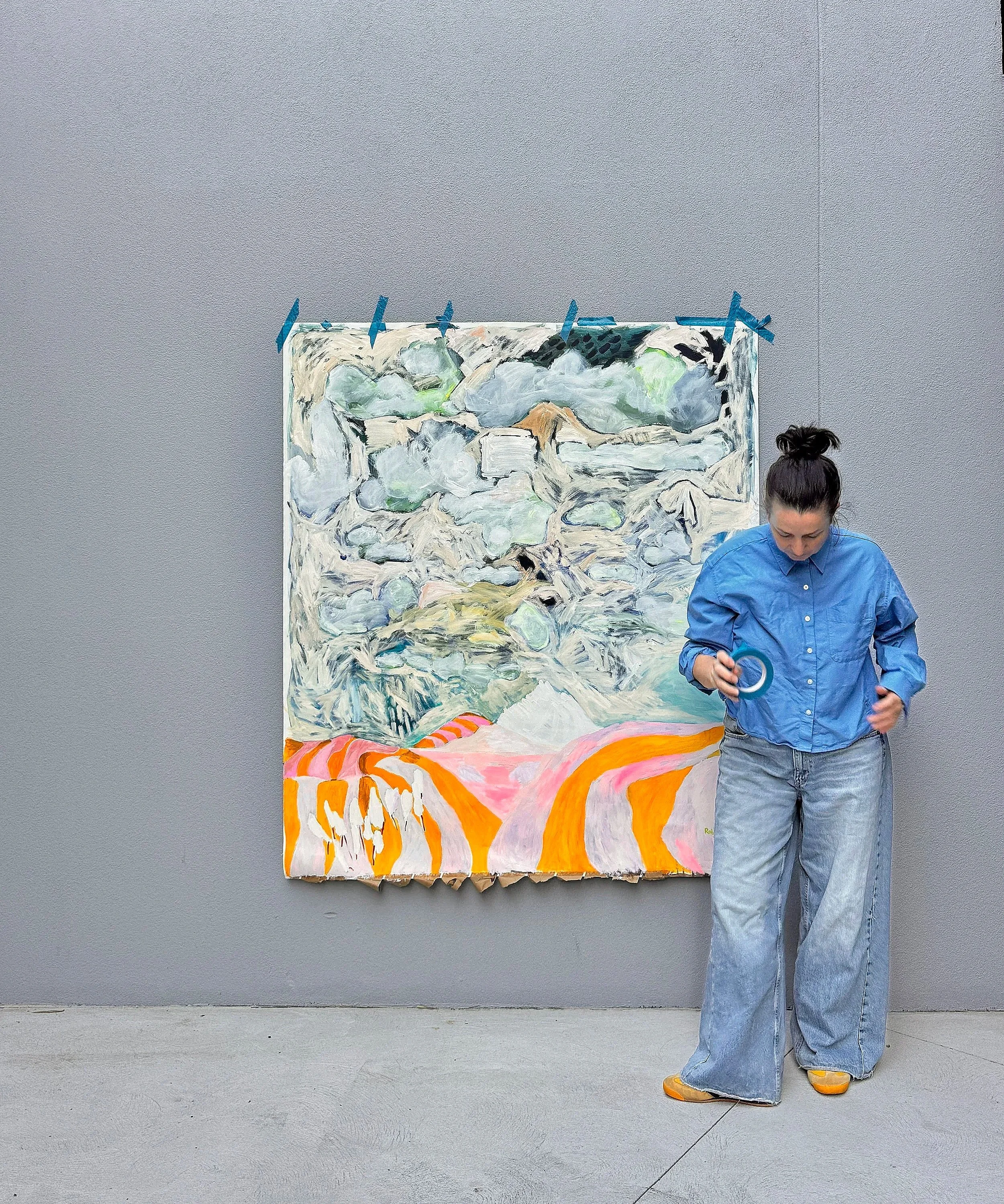 Painting on a wall with blue painter's tape and a woman in a blue shirt with big jeans, yellow shoes, and her hair in a bun in front of it.