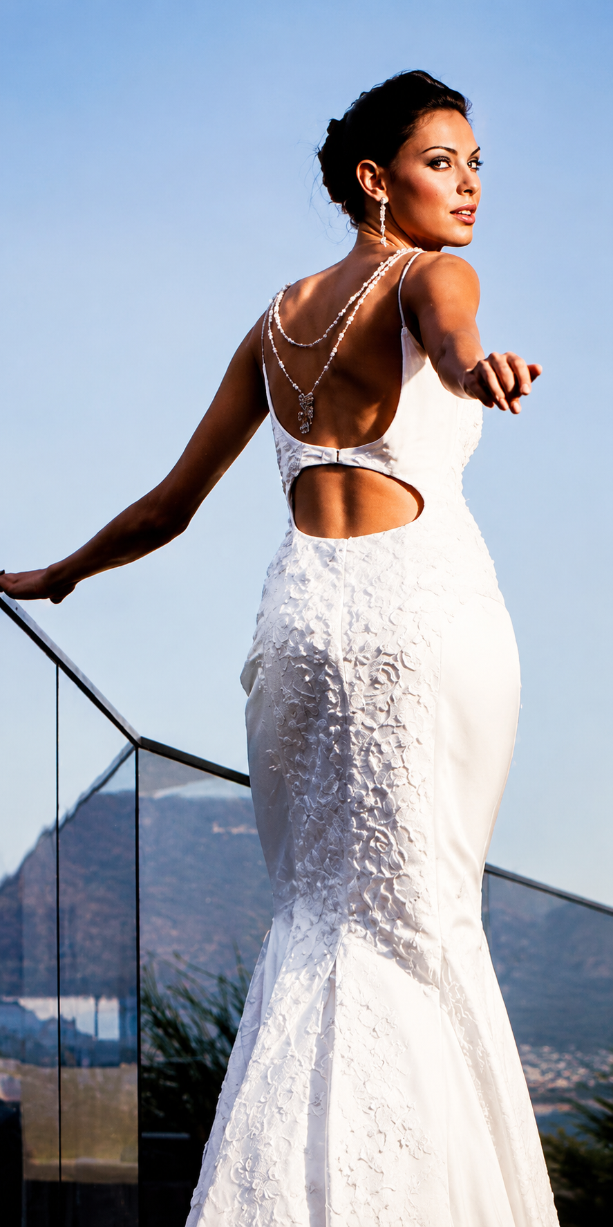 A woman in a white, textured gown with a cutout detail on the side, standing outdoors on a balcony with a view of a mountain in the background, during daytime.