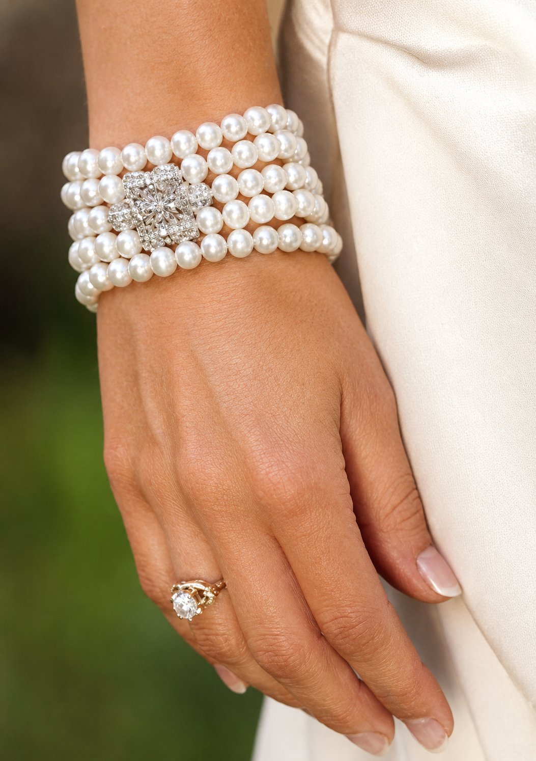 Close-up of a woman's hand wearing a multi-strand pearl bracelet with a decorative pearl and diamond clasp and a diamond engagement ring. The hand rests gently on a white fabric surface.