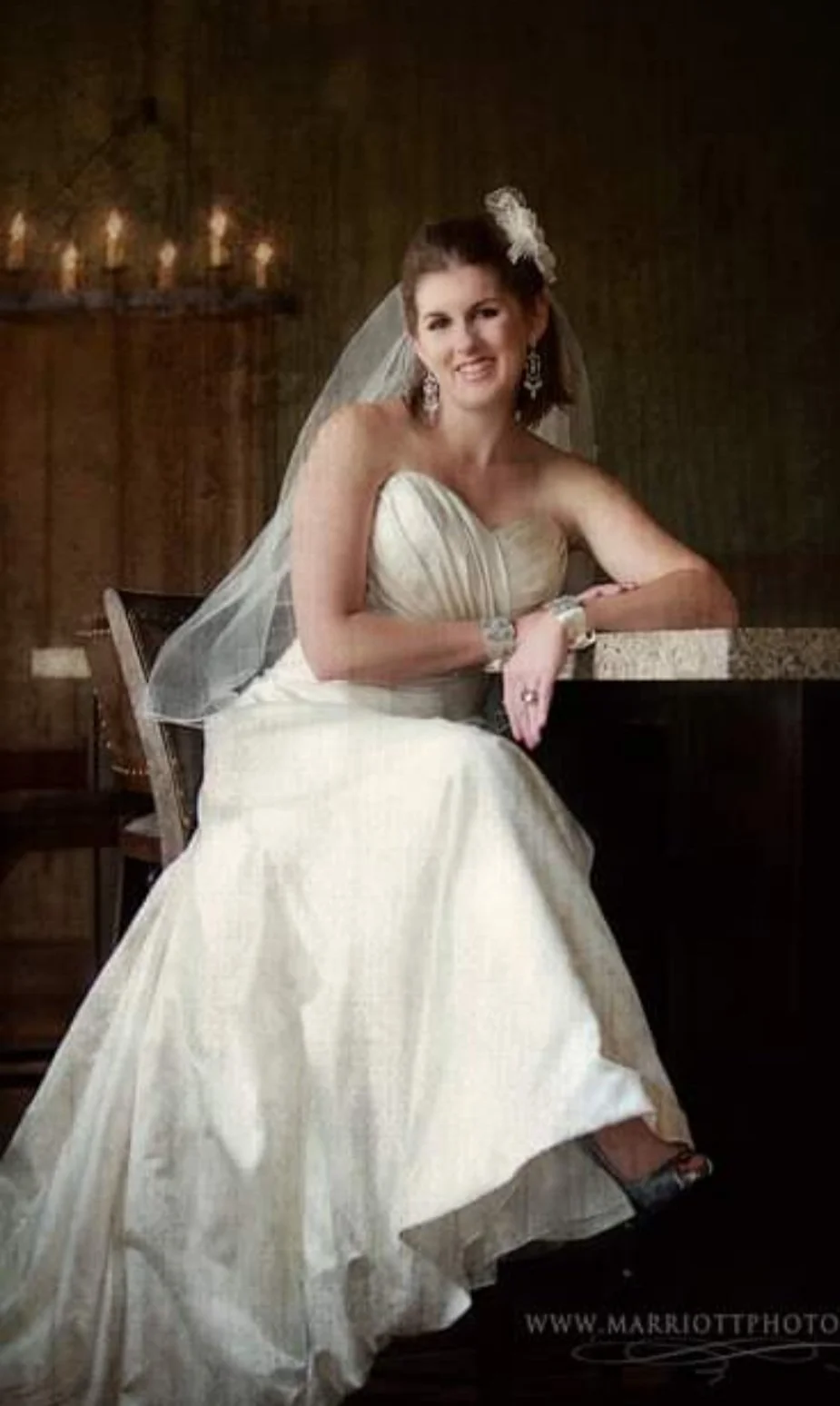 A woman in a wedding dress with a veil, sitting at a bar in a dimly-lit room with wooden walls and a row of lit candles in the background.