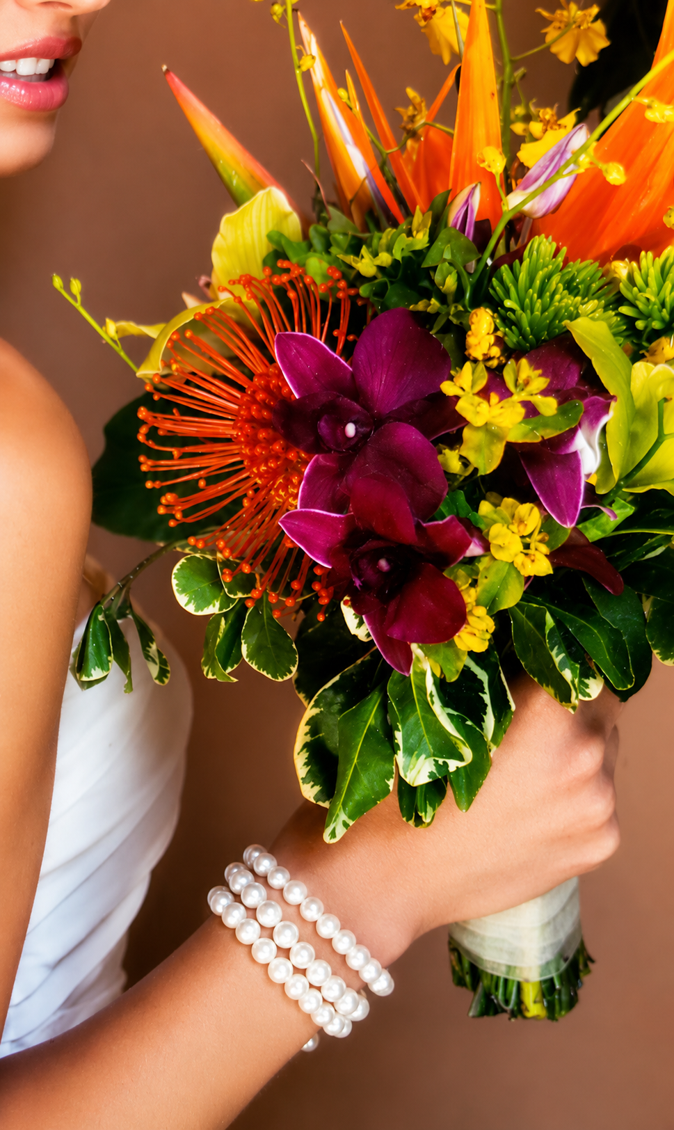 A woman in a white dress holding a colorful bouquet of tropical flowers and greenery, with pearl bracelets on her wrist.