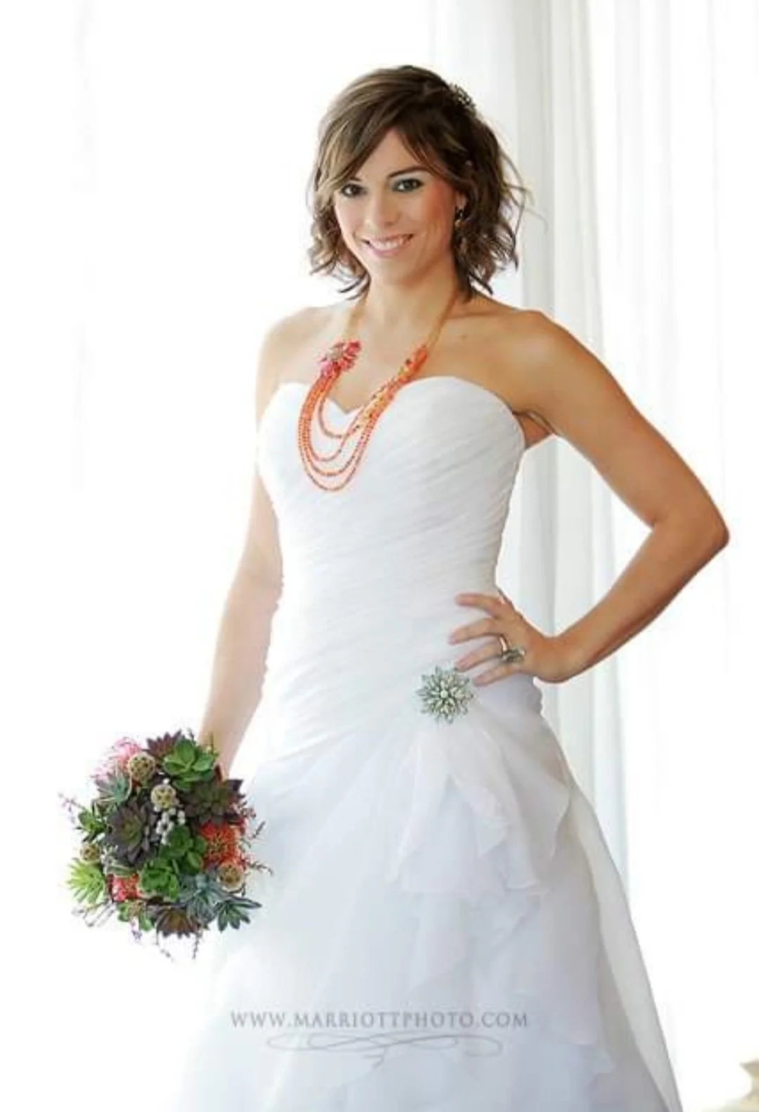 A woman in a white strapless wedding dress holding a bouquet of flowers, standing indoors with natural light.