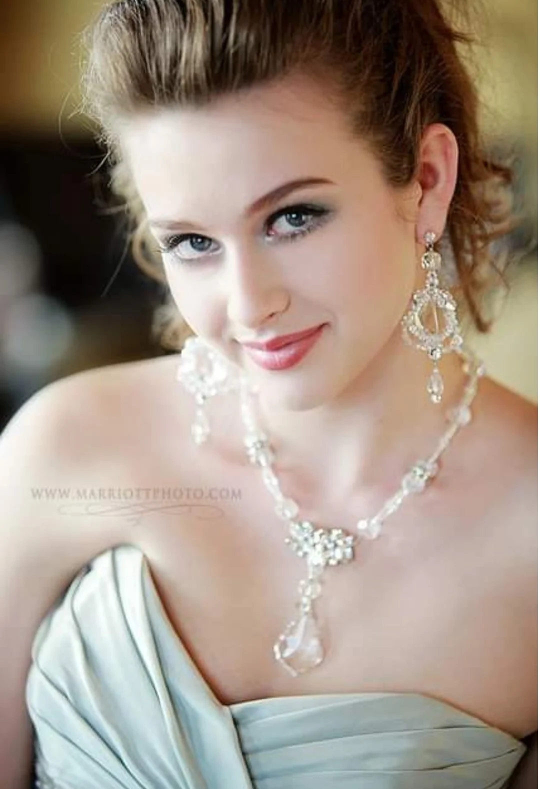 Close-up of a woman with blue eyes and red hair wearing a strapless dress and elaborate pearl and crystal jewelry, smiling at the camera.