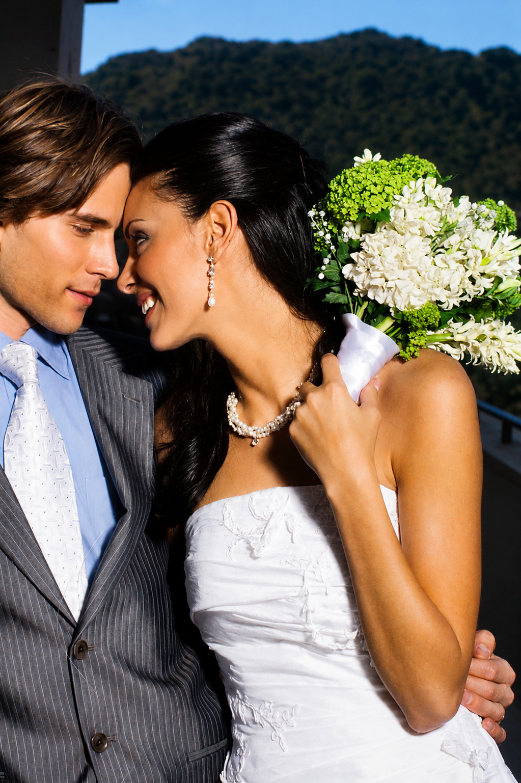 A newlywed couple sharing an intimate moment, with their foreheads touching. The bride is holding a large bouquet of white and green flowers, and they are dressed in wedding attire.