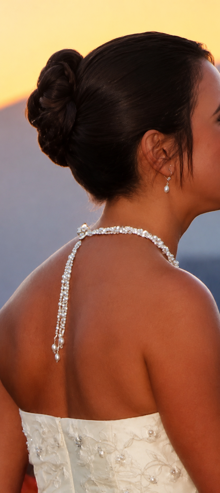 Close-up of a woman in a wedding dress with a pearl necklace and earrings, with dark hair styled in an elegant bun, against a sunset background.