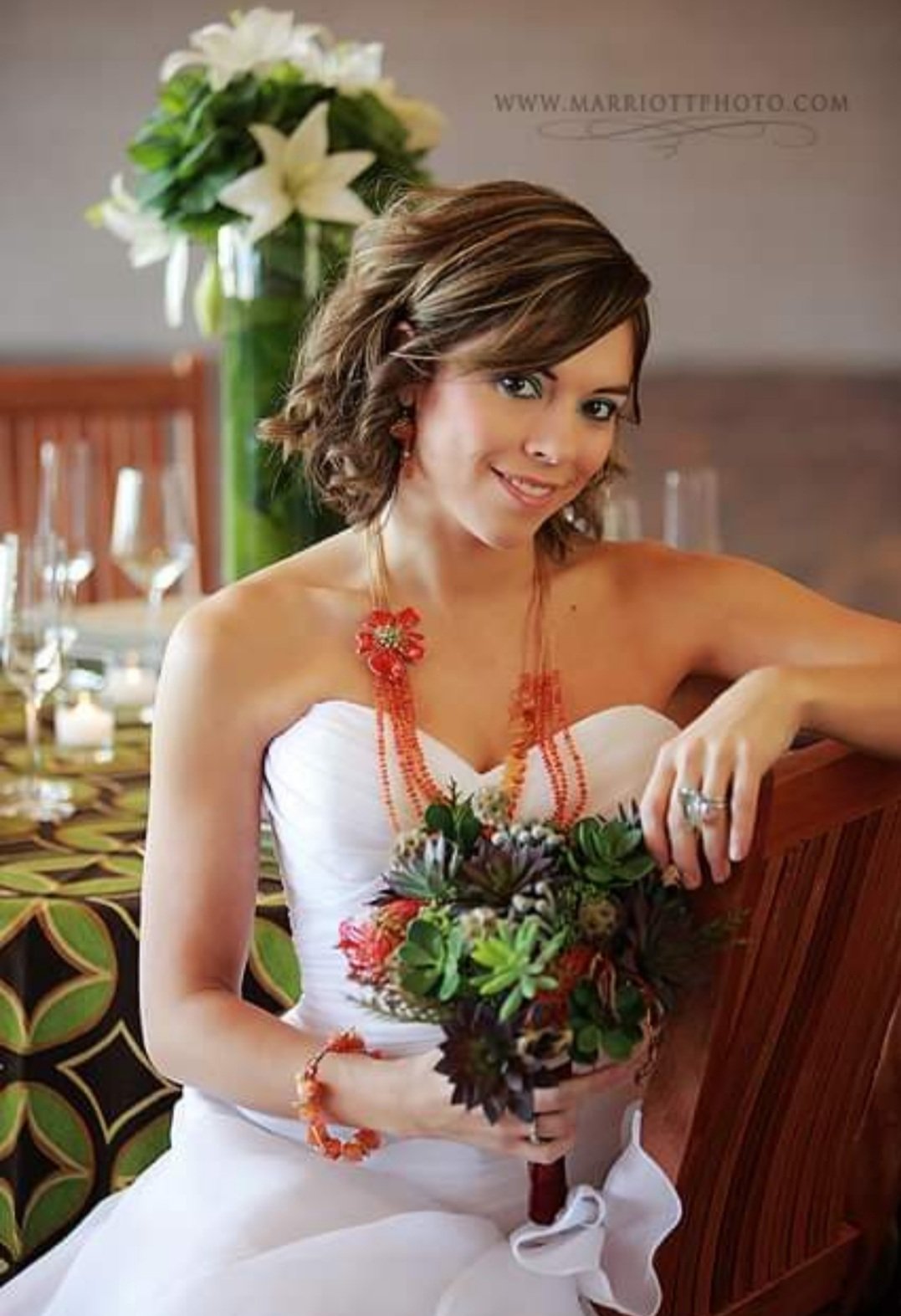 A woman in a white dress holding a small bouquet of flowers, sitting at a decorated table with a large floral arrangement in the background.