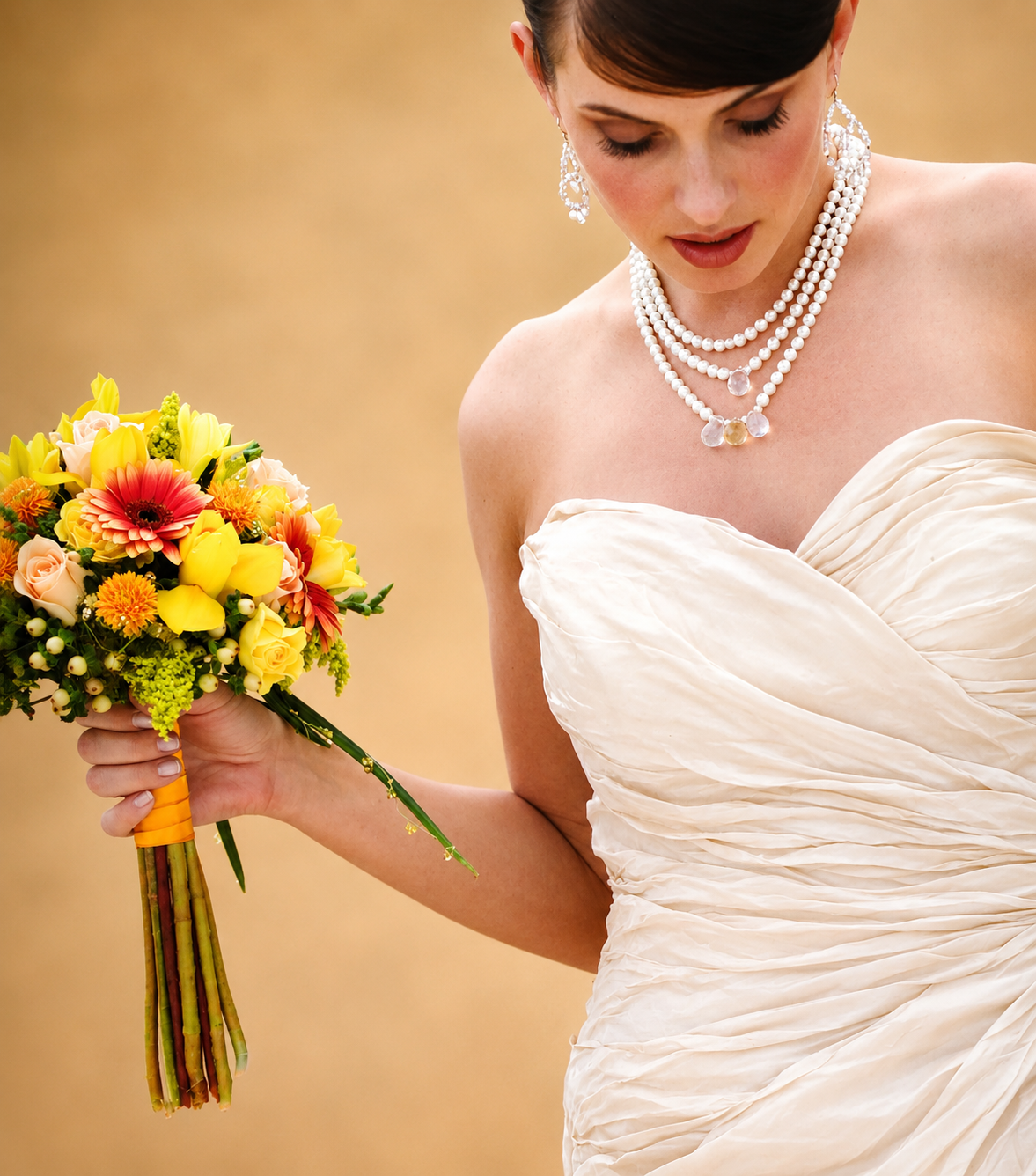 A bride holding a colorful bouquet of flowers, wearing a strapless wedding dress and pearl jewelry, with her head slightly bowed.