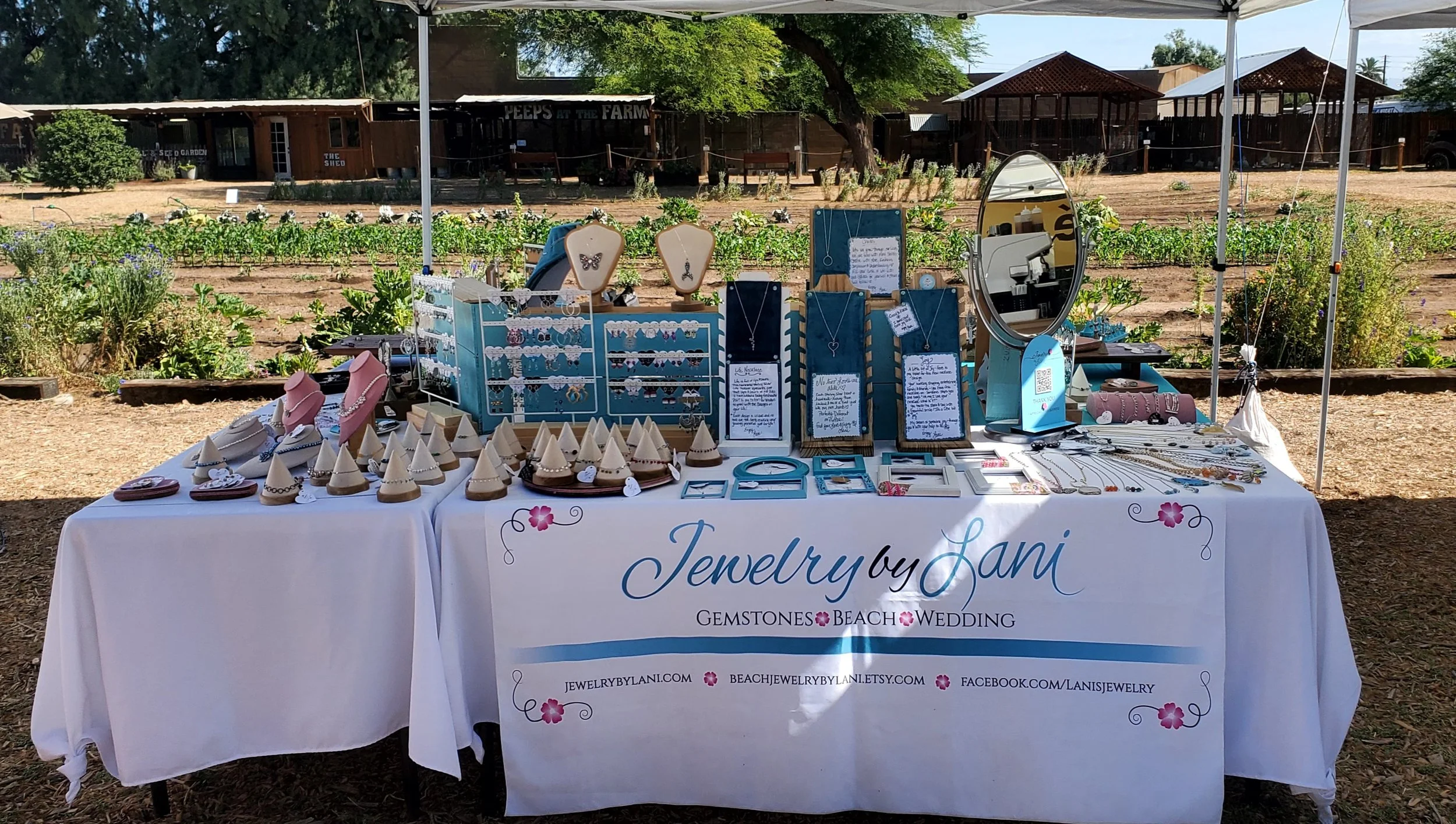 Jewelry display stand at an outdoor market, featuring necklaces, earrings, and bracelets, with a farm and garden in the background.
