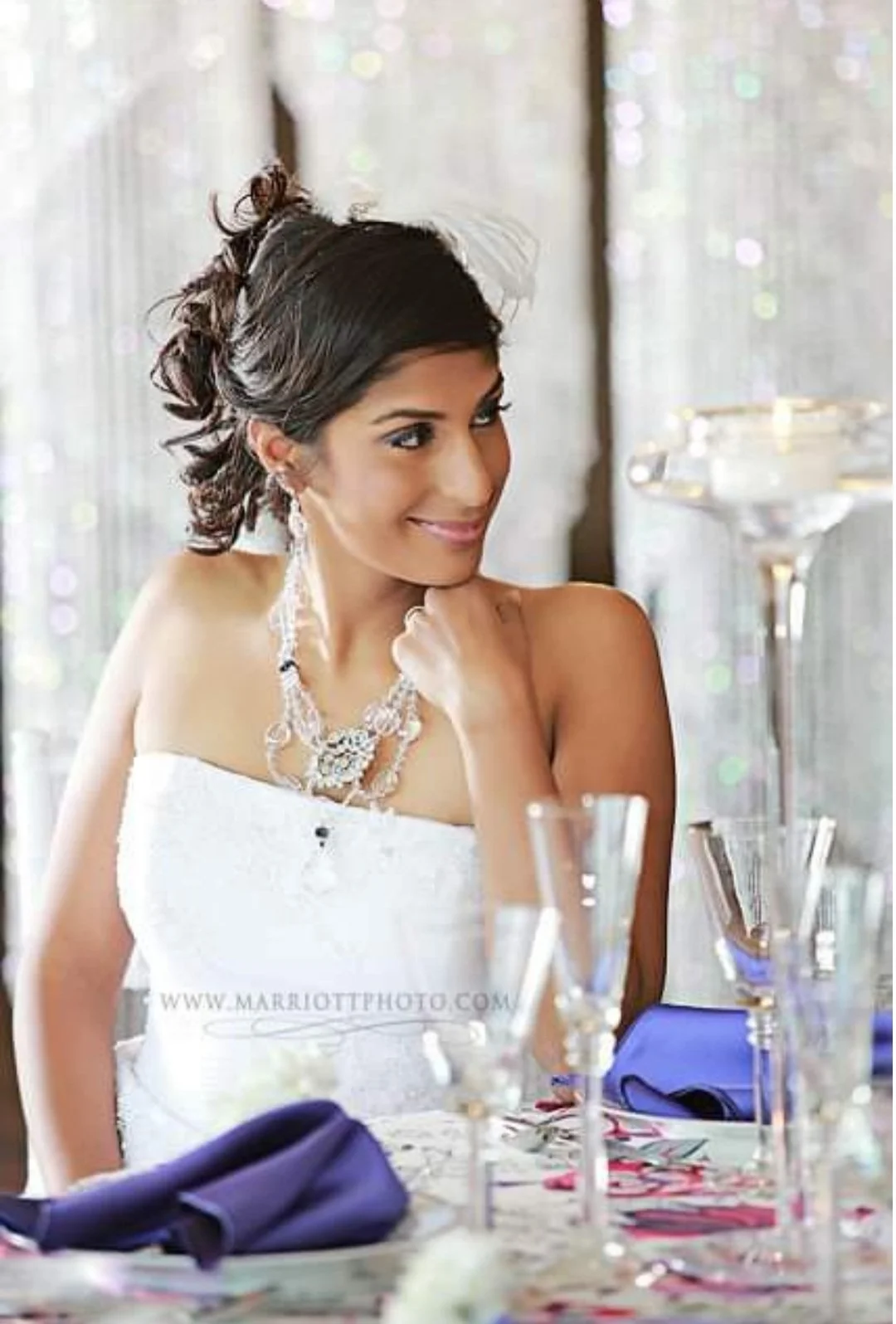 A woman in a strapless white dress, wearing an elaborate necklace, and with styled hair, sitting at a table with glassware and purple napkins. She is smiling softly and resting her chin on her hand.