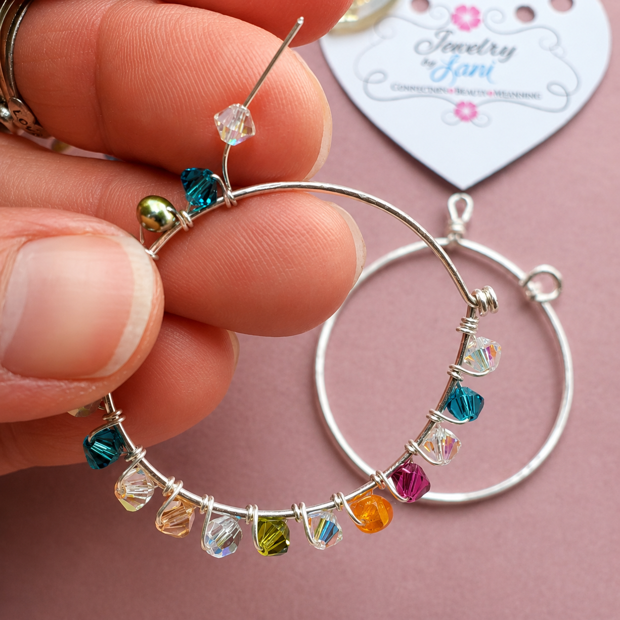 Colorful beaded hoop earrings with variously colored beads, held by a person’s fingers, with a pink background and a jewelry shop label in the background.