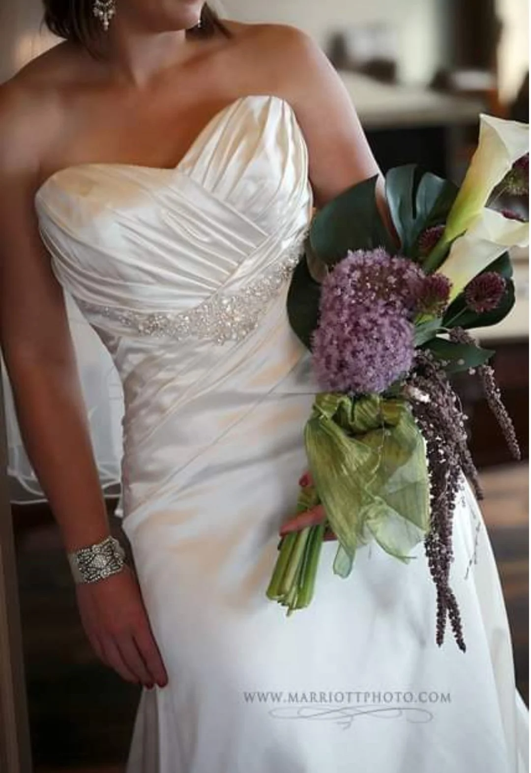 A woman wearing a white wedding dress holding a bouquet of flowers, including calla lilies, purple allium, and greenery.