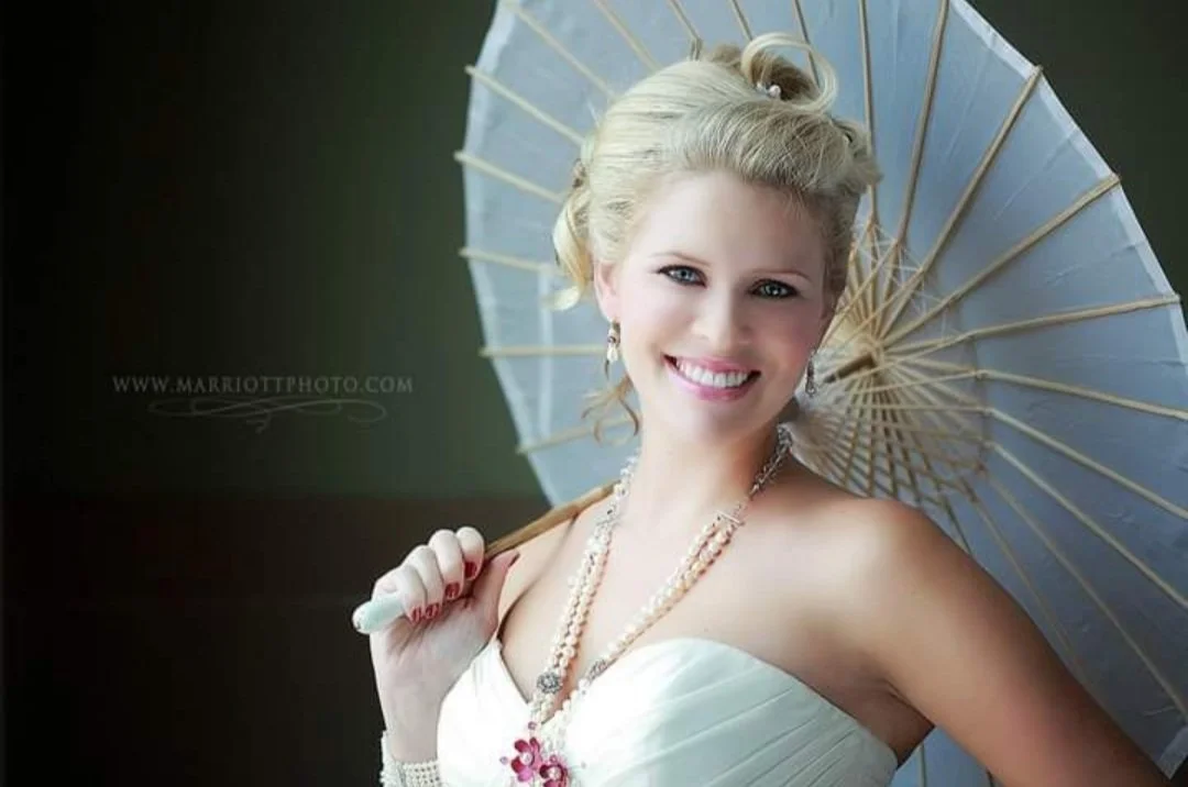 A woman with blonde hair styled in an updo, wearing a strapless white dress, pearl jewelry, and holding a light blue parasol, smiling at the camera.