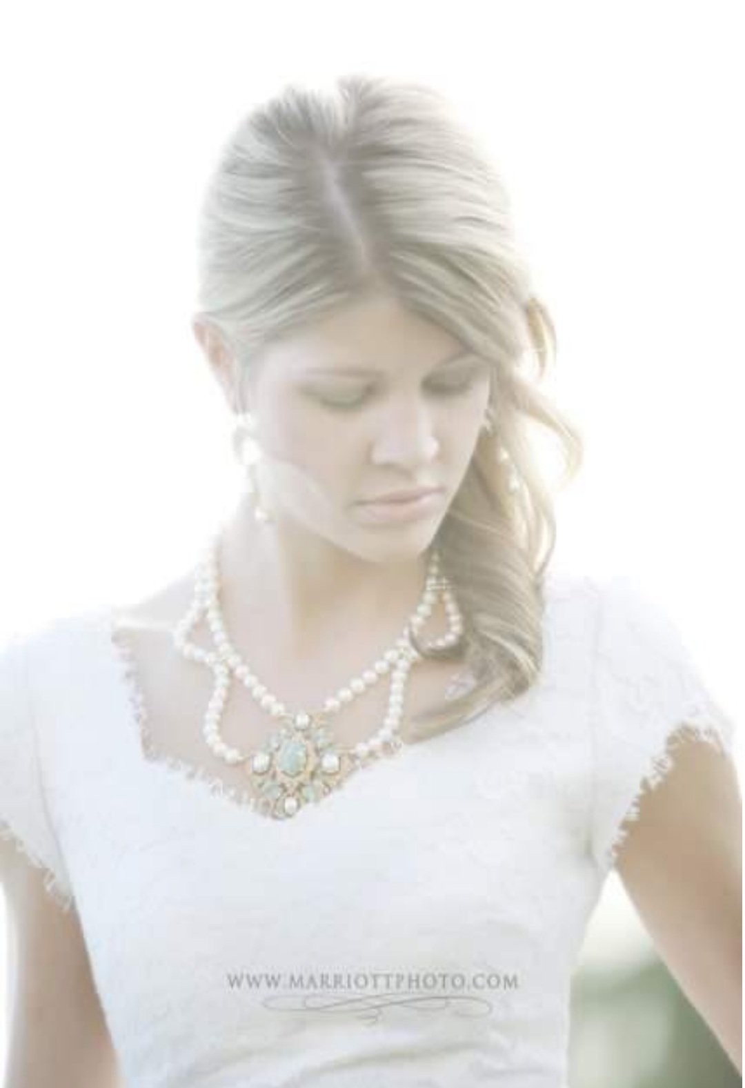 A woman with blonde hair wearing a white top and pearl necklace, looking down.n