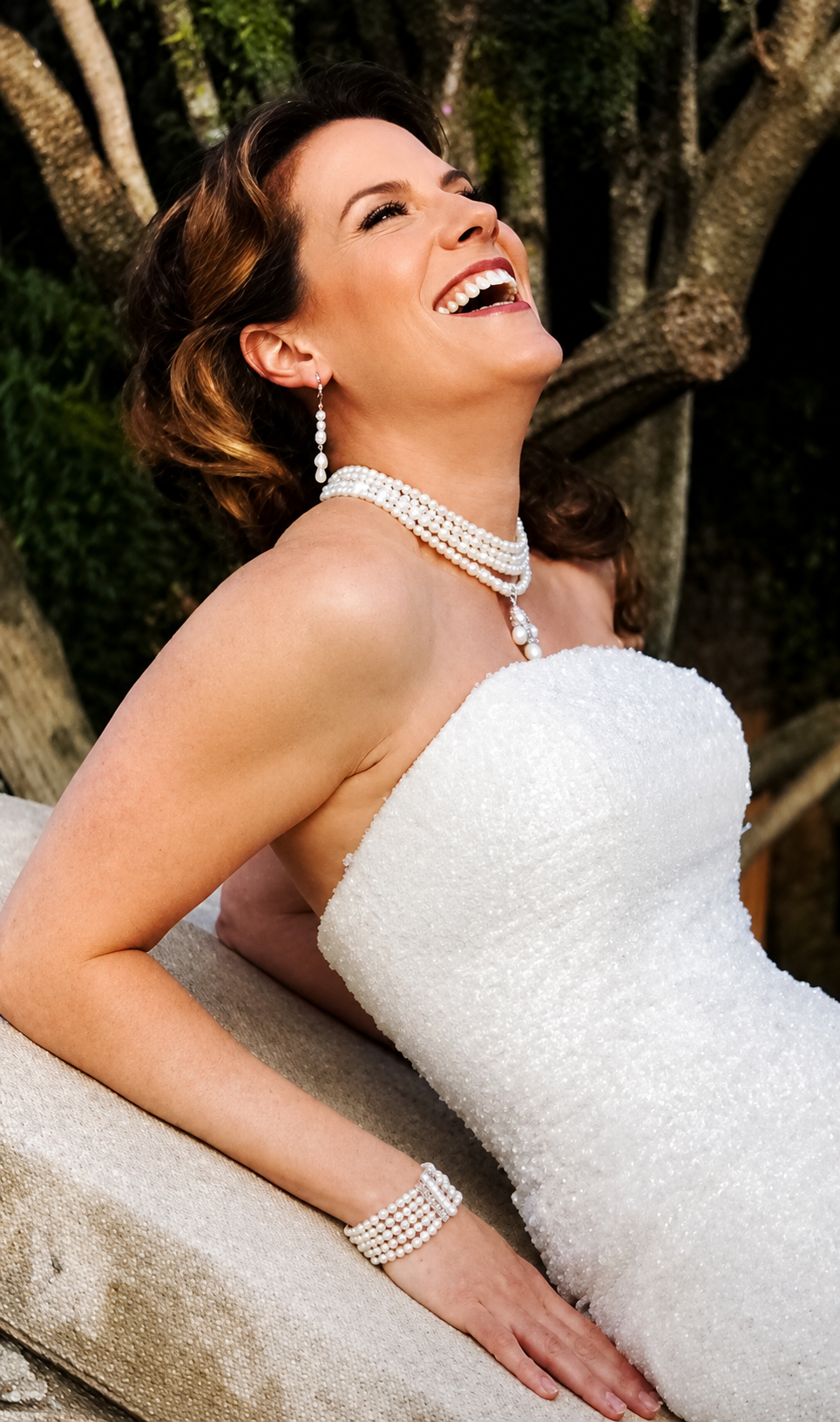 Woman in a white strapless dress, wearing pearl jewelry, smiling and laughing outdoors near trees.