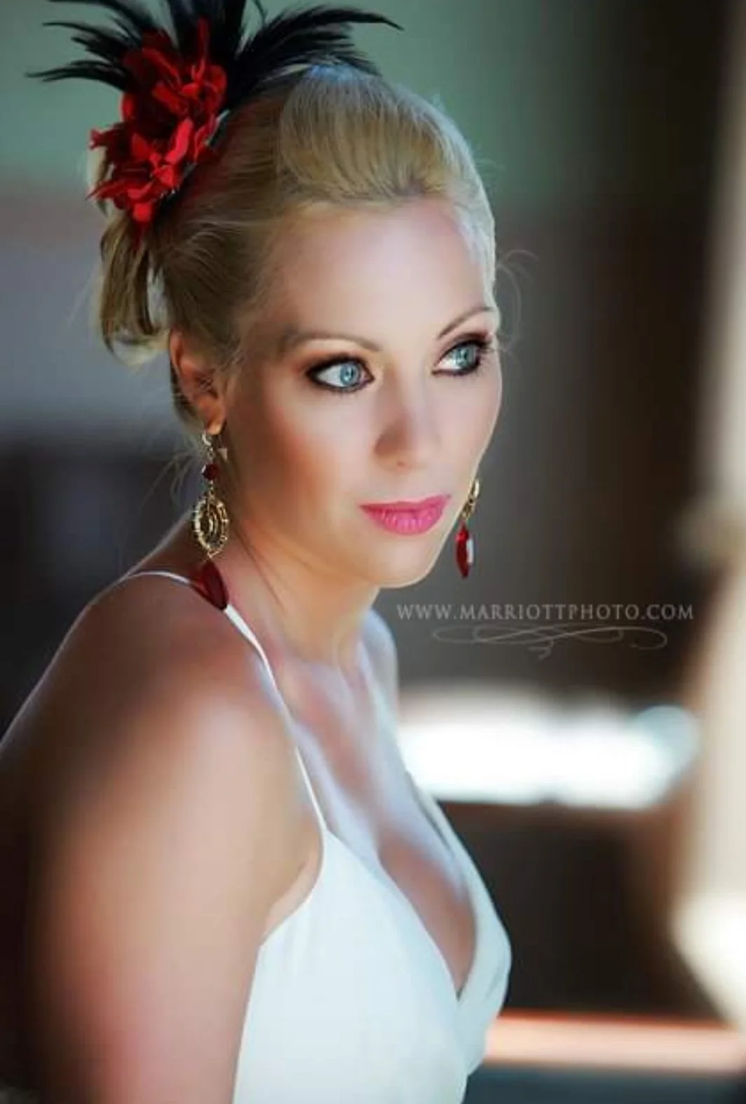 Close-up of a blonde woman with blue eyes, wearing a white dress and red jewelry, with a black and red feathered headpiece, looking off to the side.