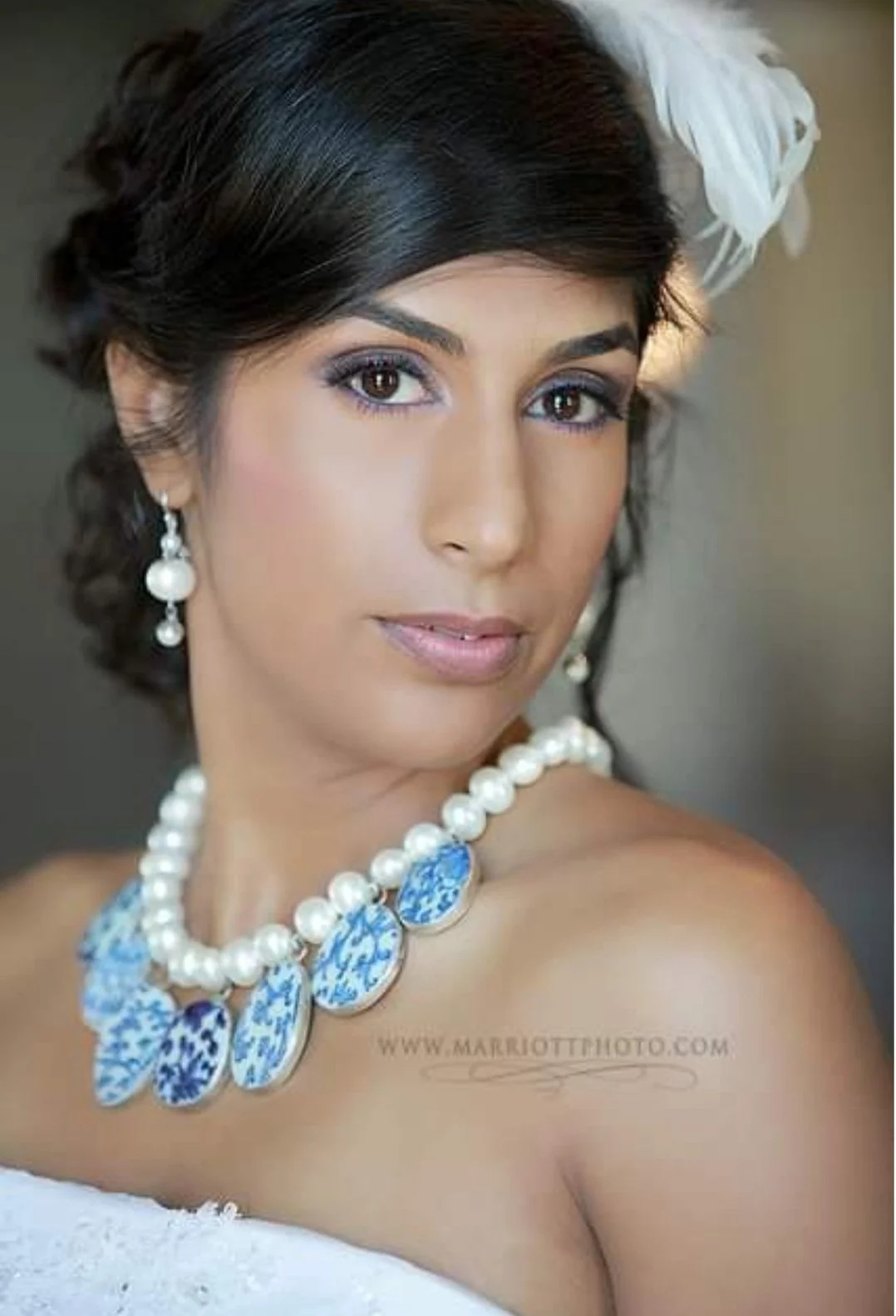 Portrait of a woman wearing pearl jewelry and a white flower in her dark hair, with a soft, neutral background.