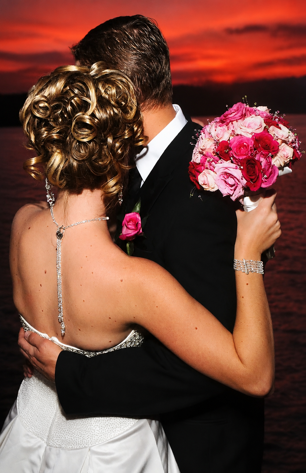 A bride and groom embracing, with the bride holding a bouquet of pink and red roses during sunset by the water.