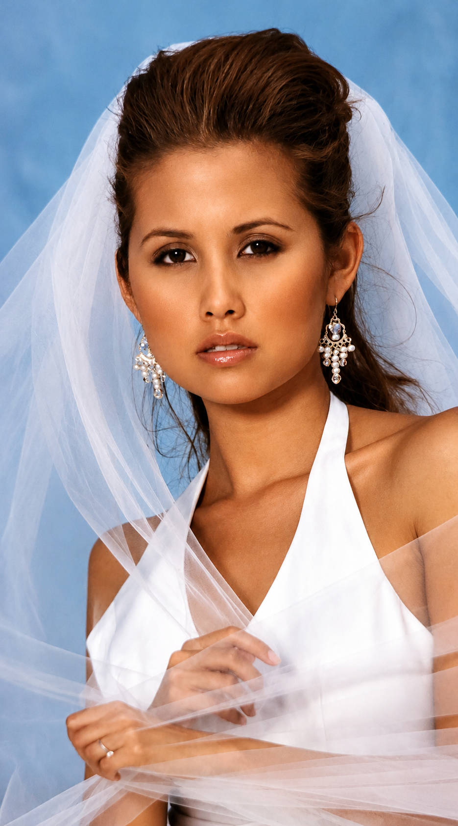 A woman with brown hair styled in a bouffant, wearing a white wedding dress and veil, with jewelry, against a light blue background.