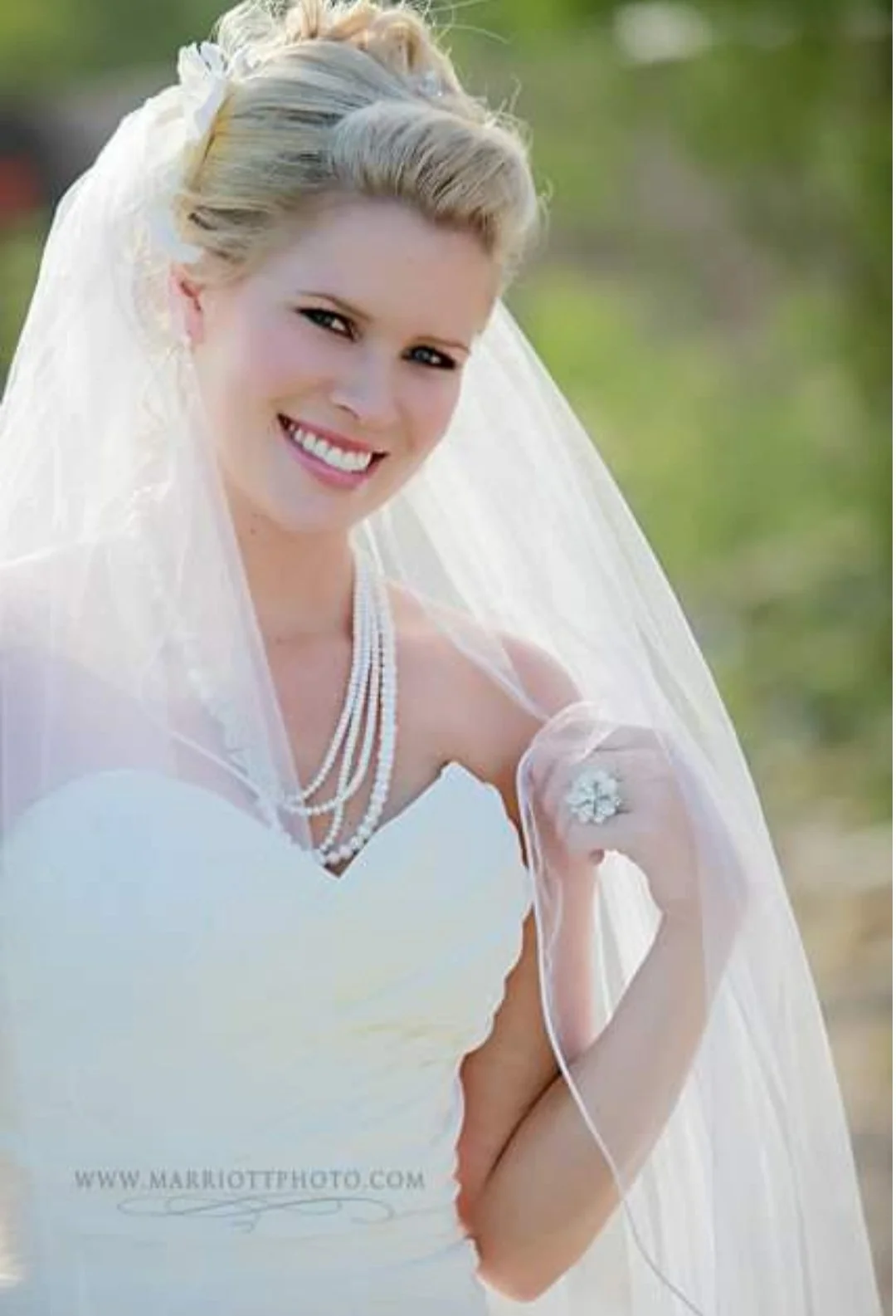 A smiling bride in a white wedding dress, with a veil, pearl necklace, and floral headpiece, outdoors on a sunny day.
