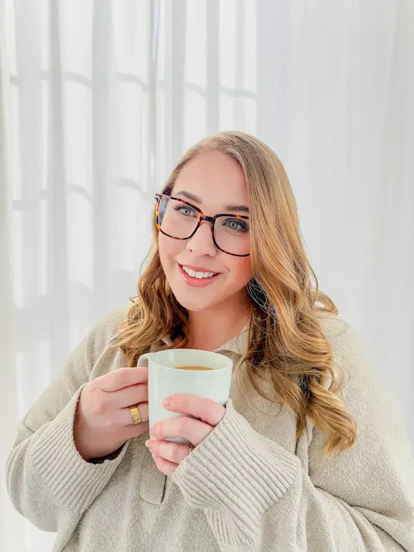 A woman with long wavy hair, glasses, and a beige sweater holds a white mug with both hands and smiles, standing in front of a sheer white curtain.