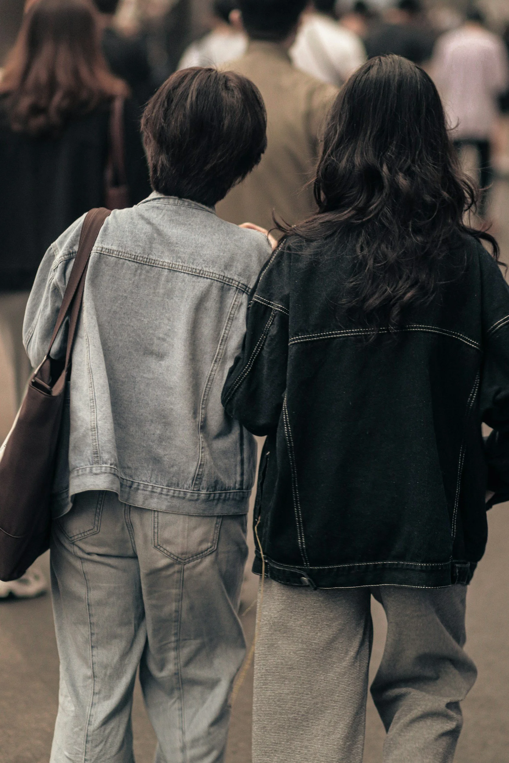 Two people walking in an indoor crowd, seen from behind. The person on the left has short dark hair and is wearing a light denim jacket and pants. The person on the right has long dark wavy hair and is dressed in a black jacket and gray pants.