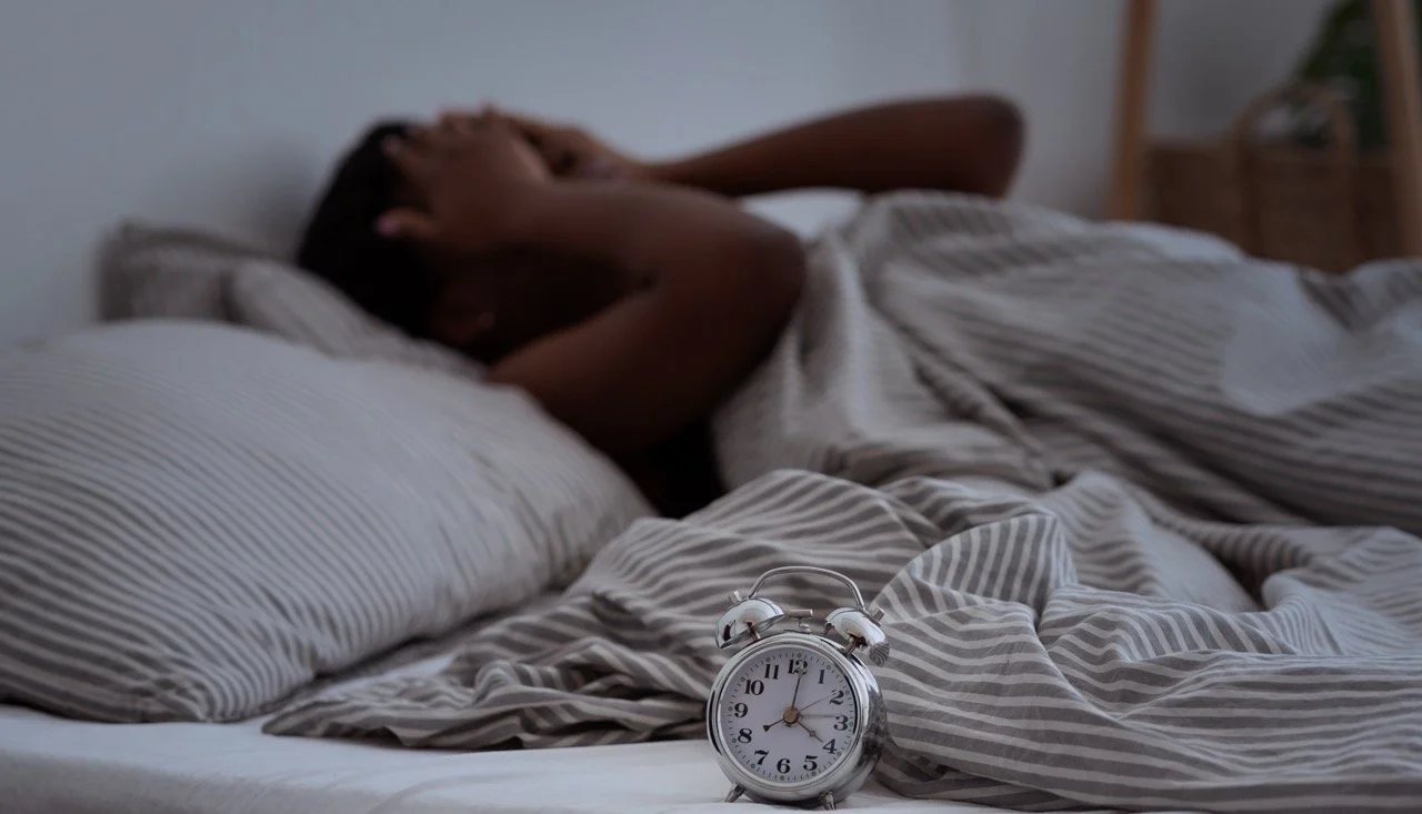 A person lying in bed with hands over their face, and an alarm clock showing 7:10 in the foreground.