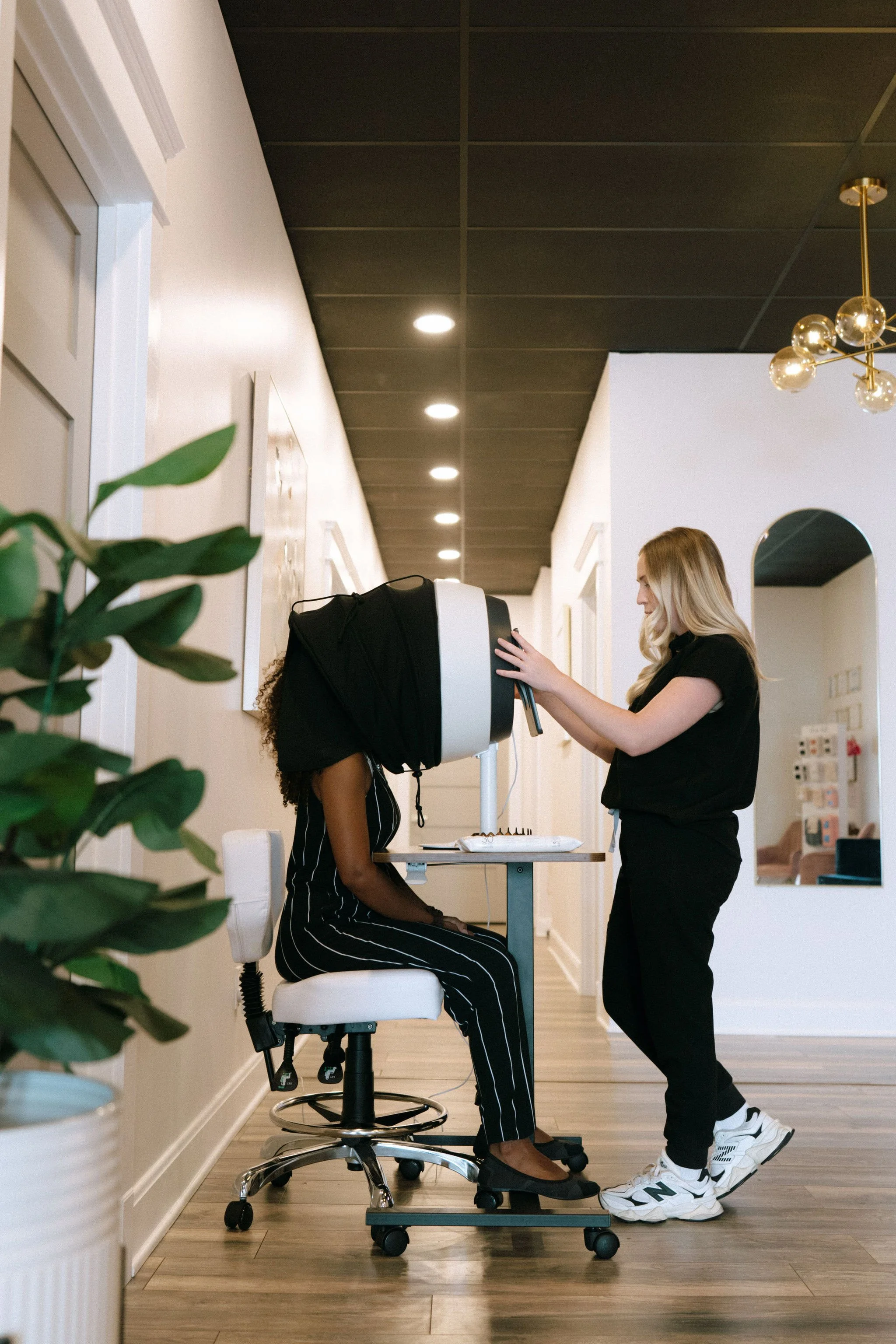 A woman sitting on a stool at a skincare consultation, with another woman standing and adjusting a device on her head to get scans of her face.