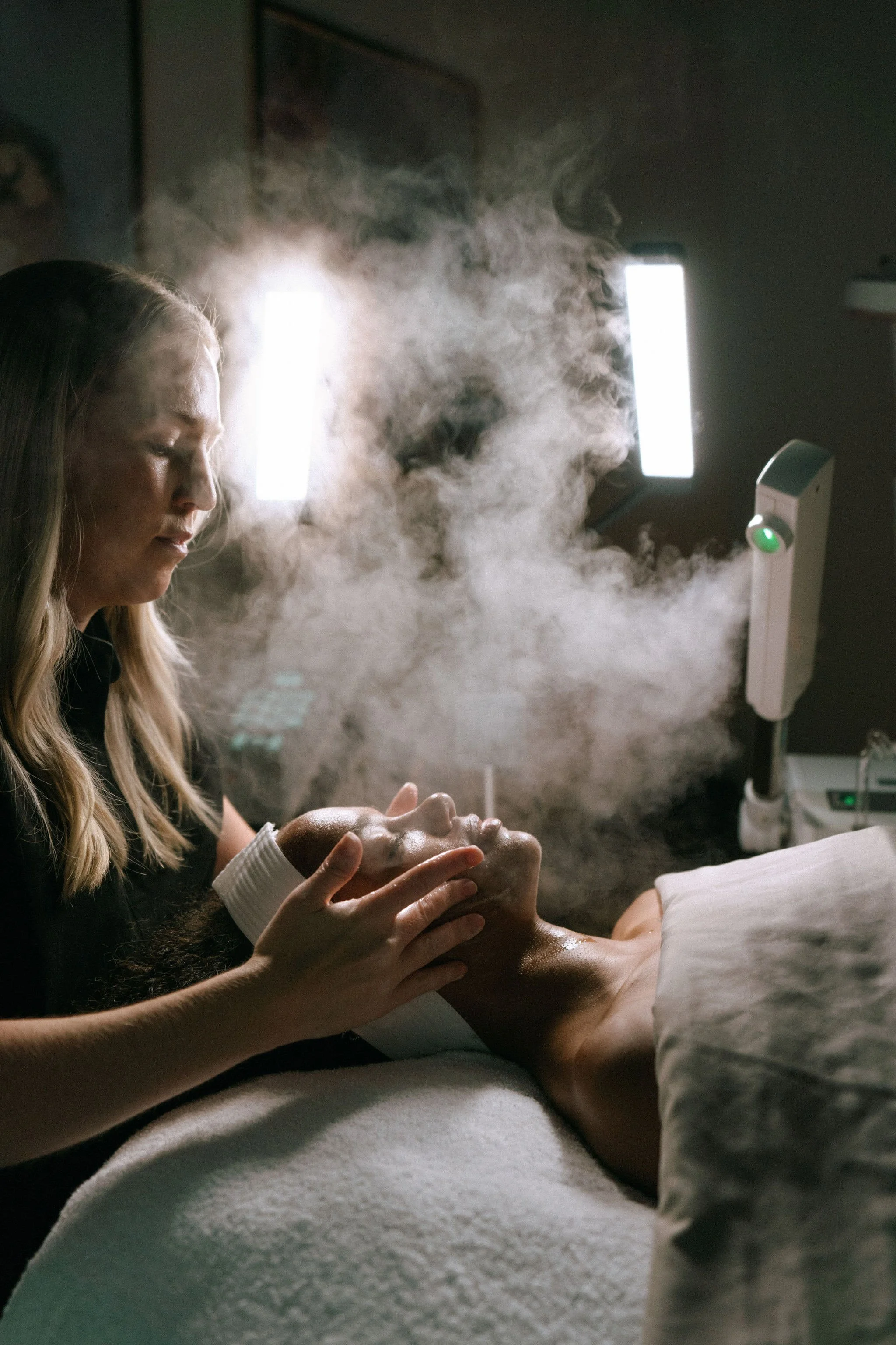 A woman performing a facial treatment on a person lying on a bed, with steam or vapor around them in a dimly lit room.