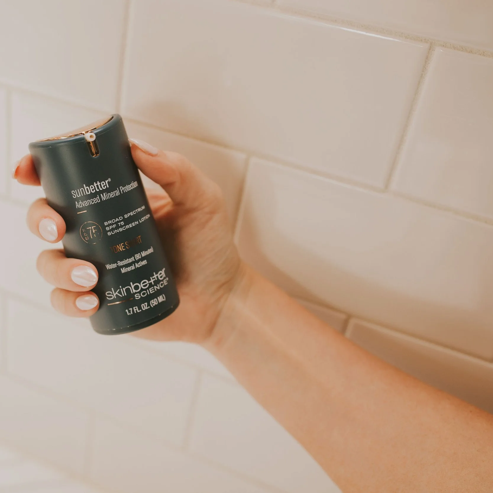 A person holds a container of Sunbetter Advanced Mineral Protection sunscreen lotion in front of a beige tiled wall.