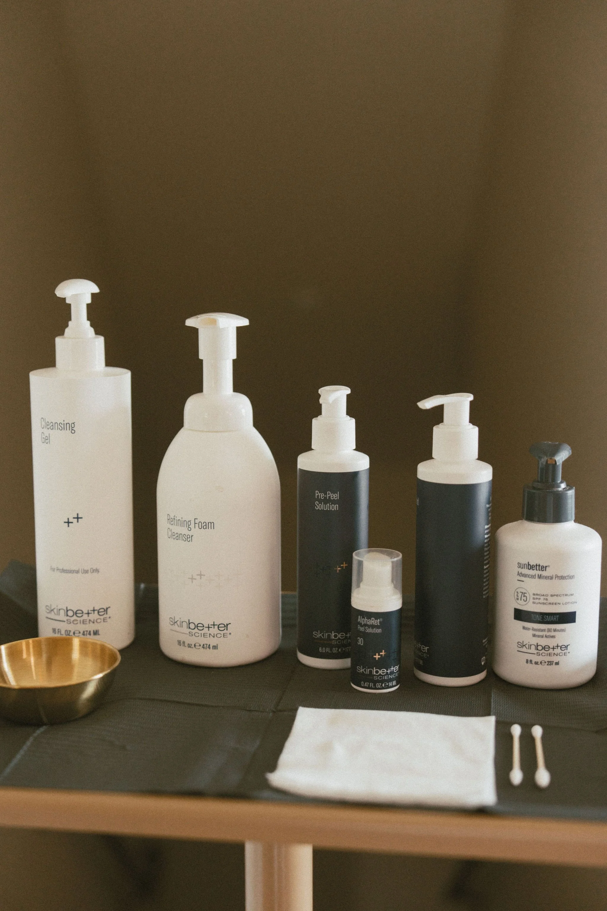 Assorted skincare products, including cleansing gel, refining foam cleanser, pre-peel solution, and sunscreen, are displayed on a black surface with a small gold bowl and cotton swabs nearby.