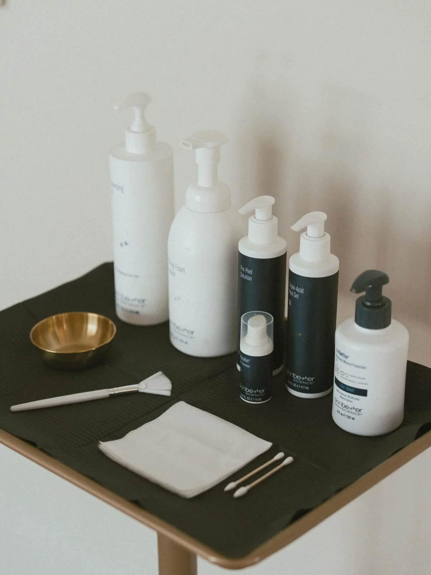 A black table with skincare bottles, a white brush, a gold dish, and cotton swabs, set against a beige wall.