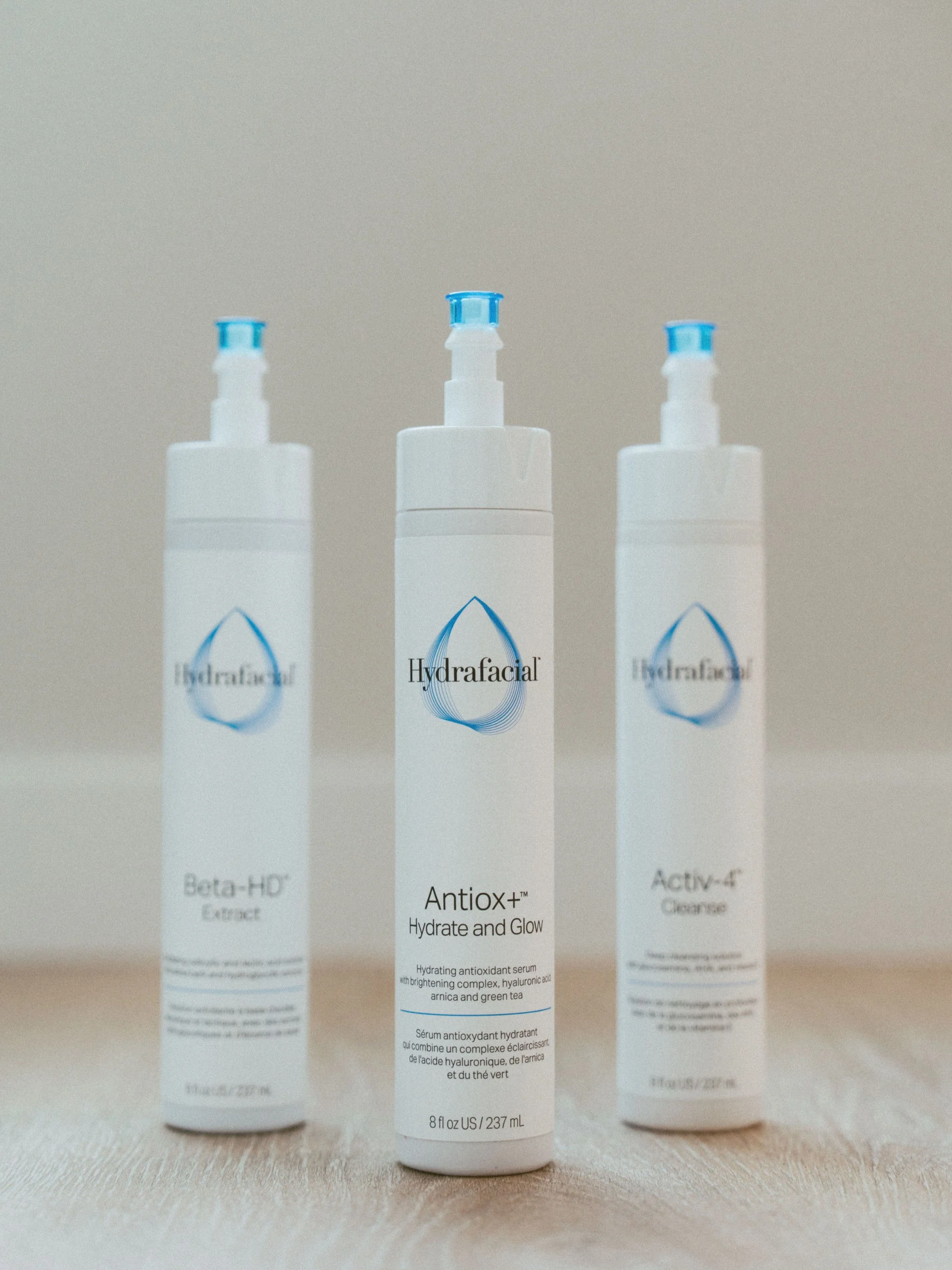 Three white bottles of skincare serums labeled Hydrafacial, with the middle one named Antiox+ Hydrate and Glow, placed on a wooden surface with a plain background.