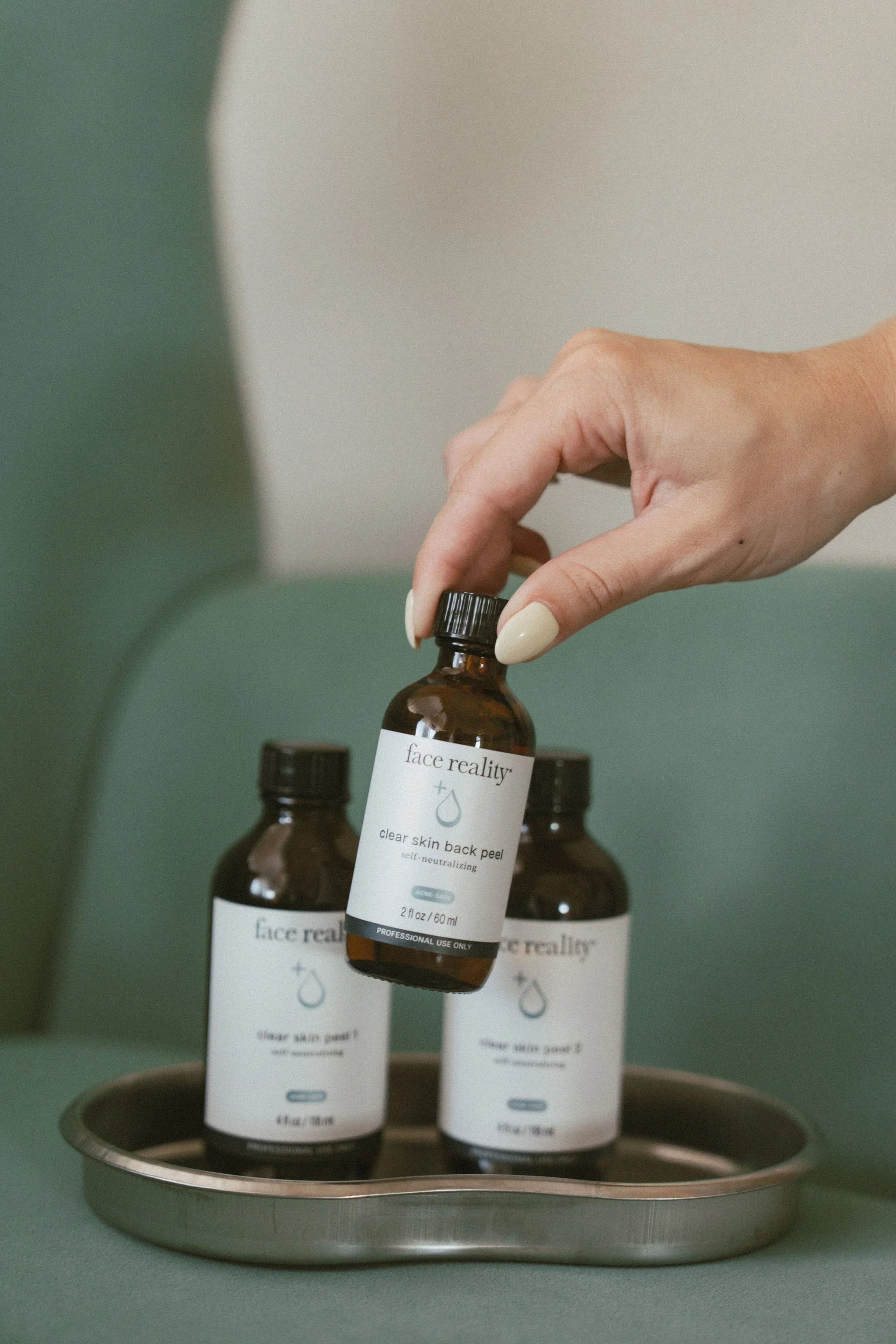 A hand reaching to pick up a brown glass bottle with a white label reading "face reality + clear skin back peel" on a metal tray, with other similar bottles in the background.