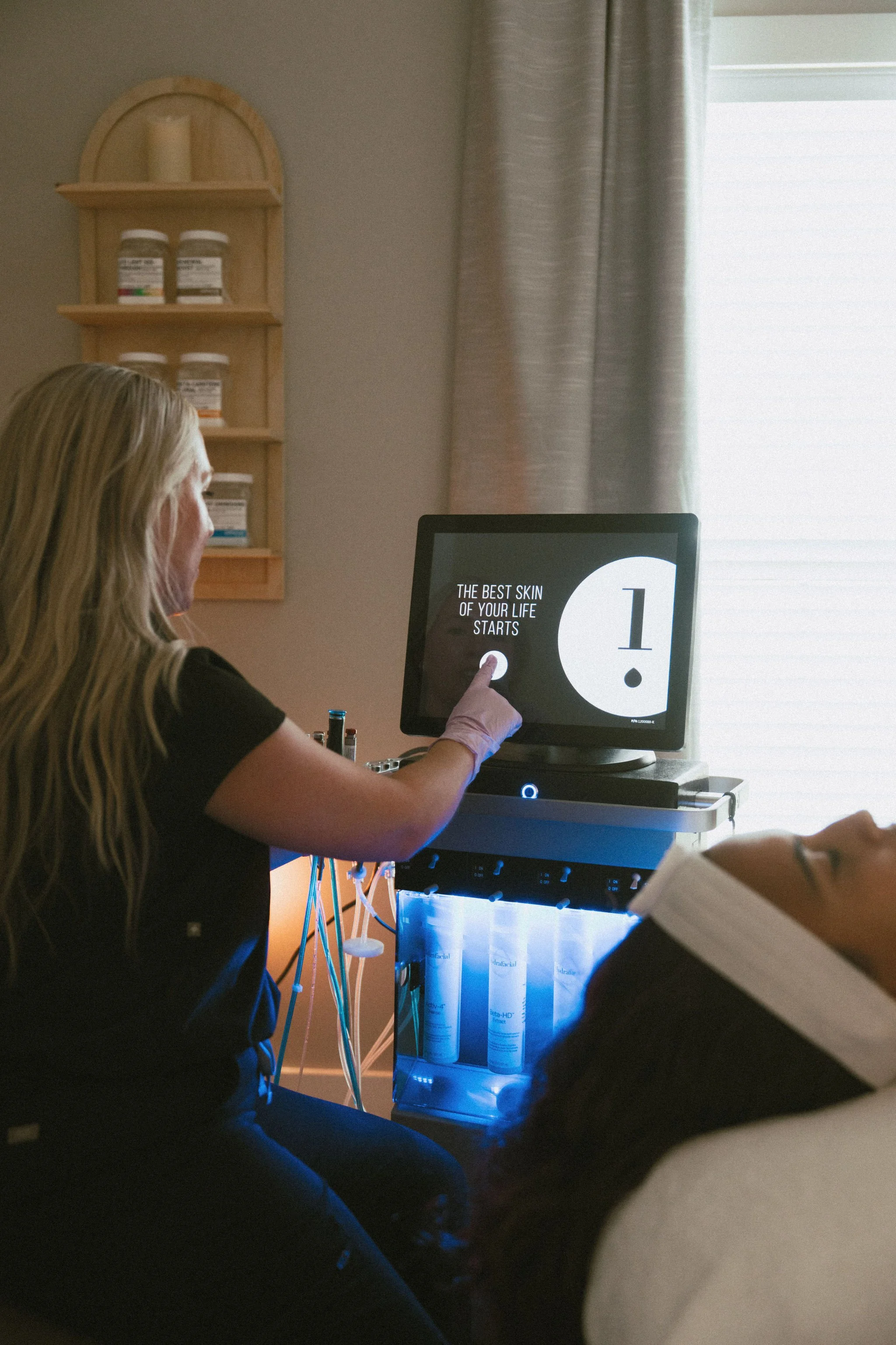 A woman with long blond hair and wearing black scrubs, pointing to a computer screen that says 'The best skin of your life starts.' In the background, there is a wooden shelf with bottles, a window with blinds, and a skincare product display with bottles glowing blue.