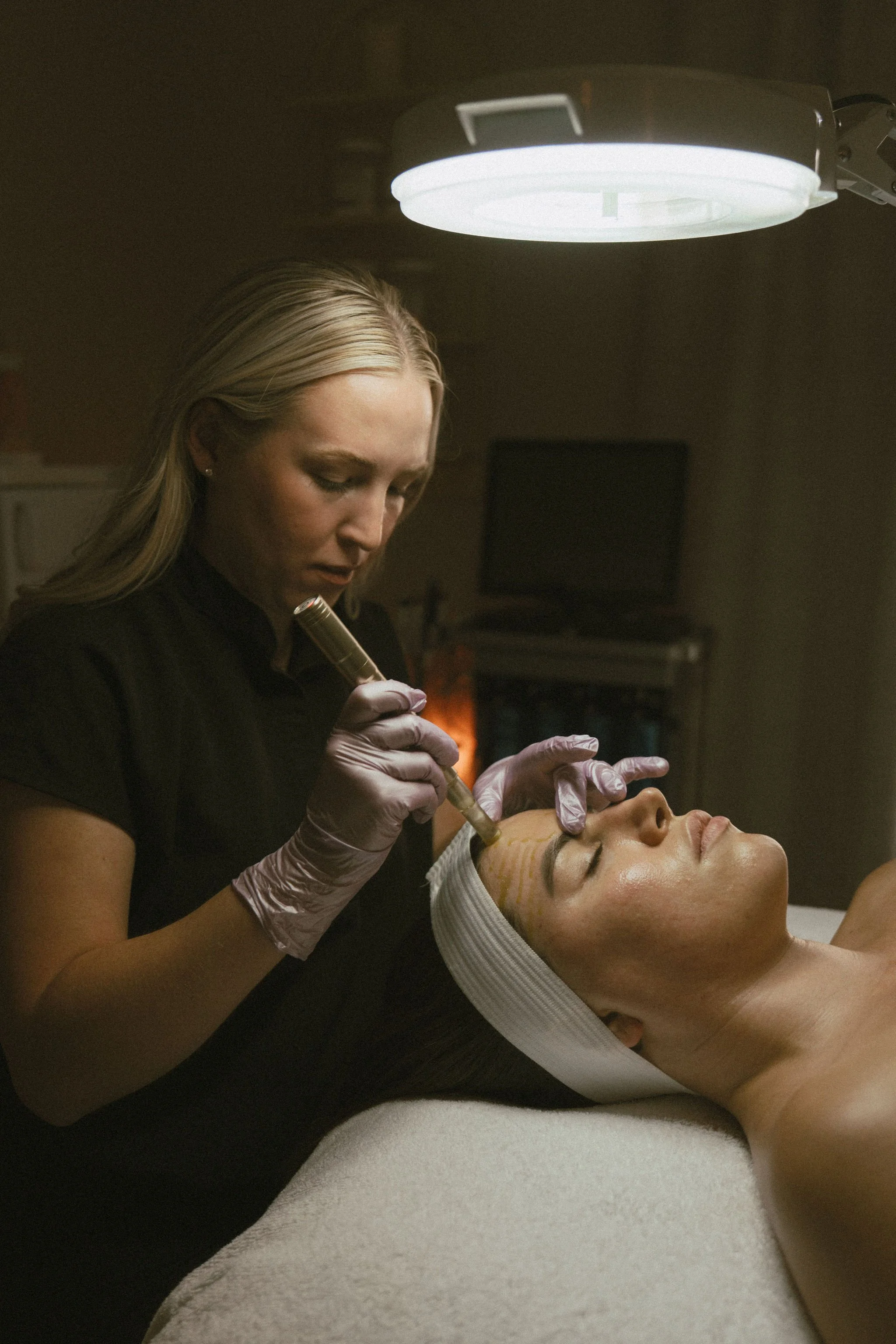 A woman receiving a cosmetic facial treatment while lying on a table, with a professional woman in gloves applying a procedure to her face, in a spa or clinic setting.