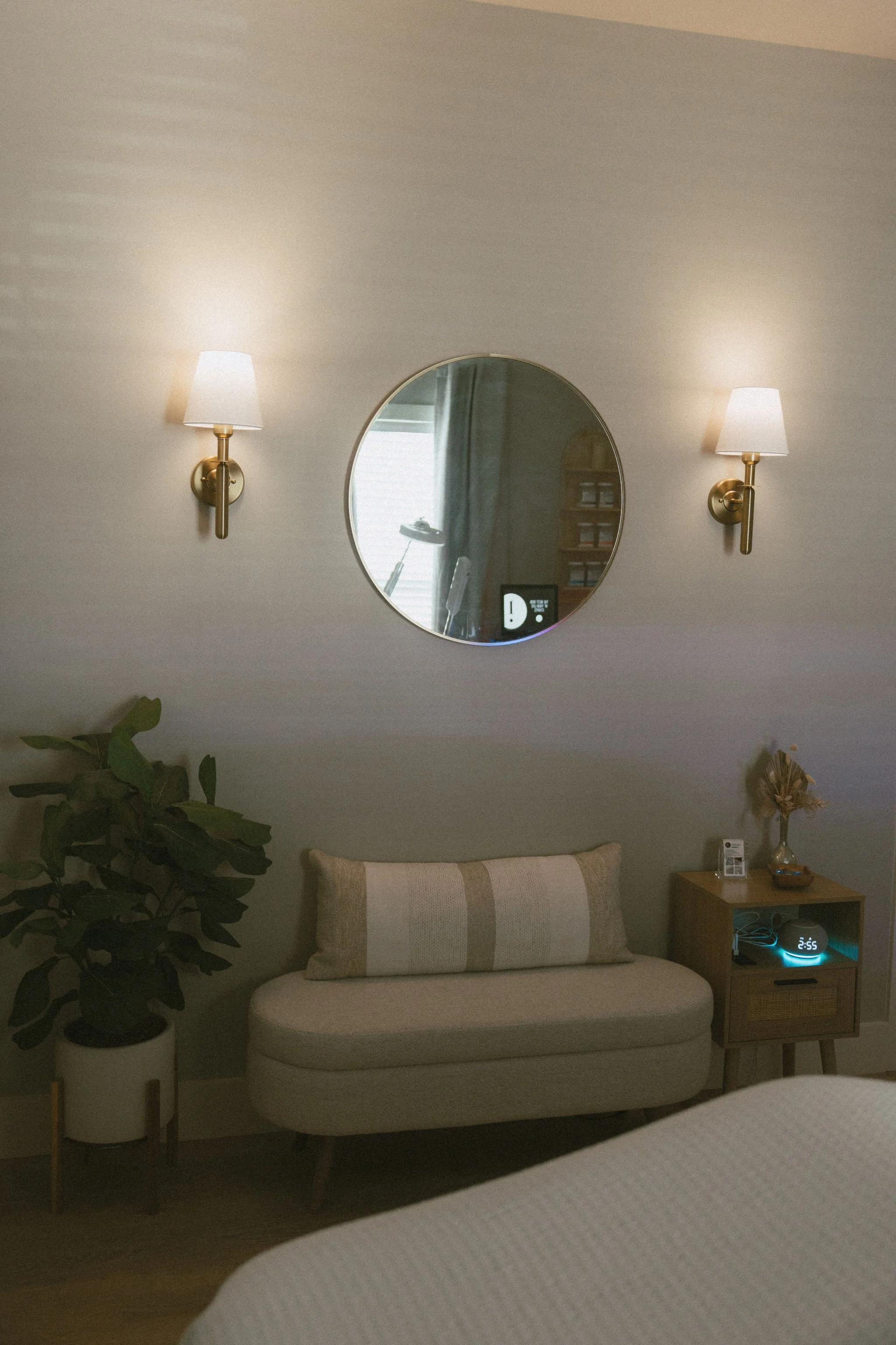 corner featuring a beige bench with a matching pillow, a wooden side table with a digital clock, small decorative items, a tall potted plant, a circular mirror on the wall, and two wall-mounted lamps with white shades, all with soft ambient lighting.