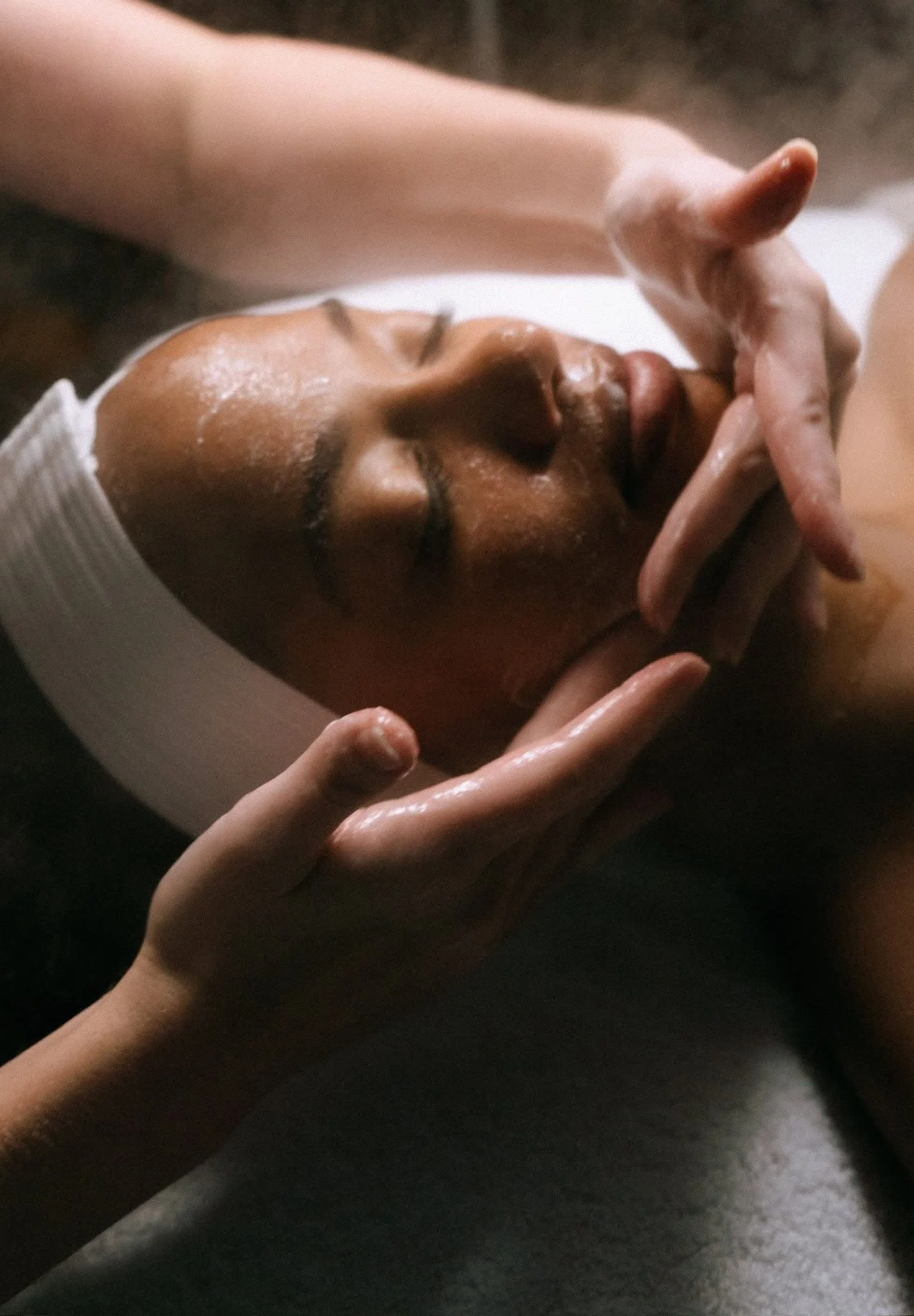 Close-up of a person lying down, with their eyes closed, receiving a facial massage from another person's hands in a spa setting.