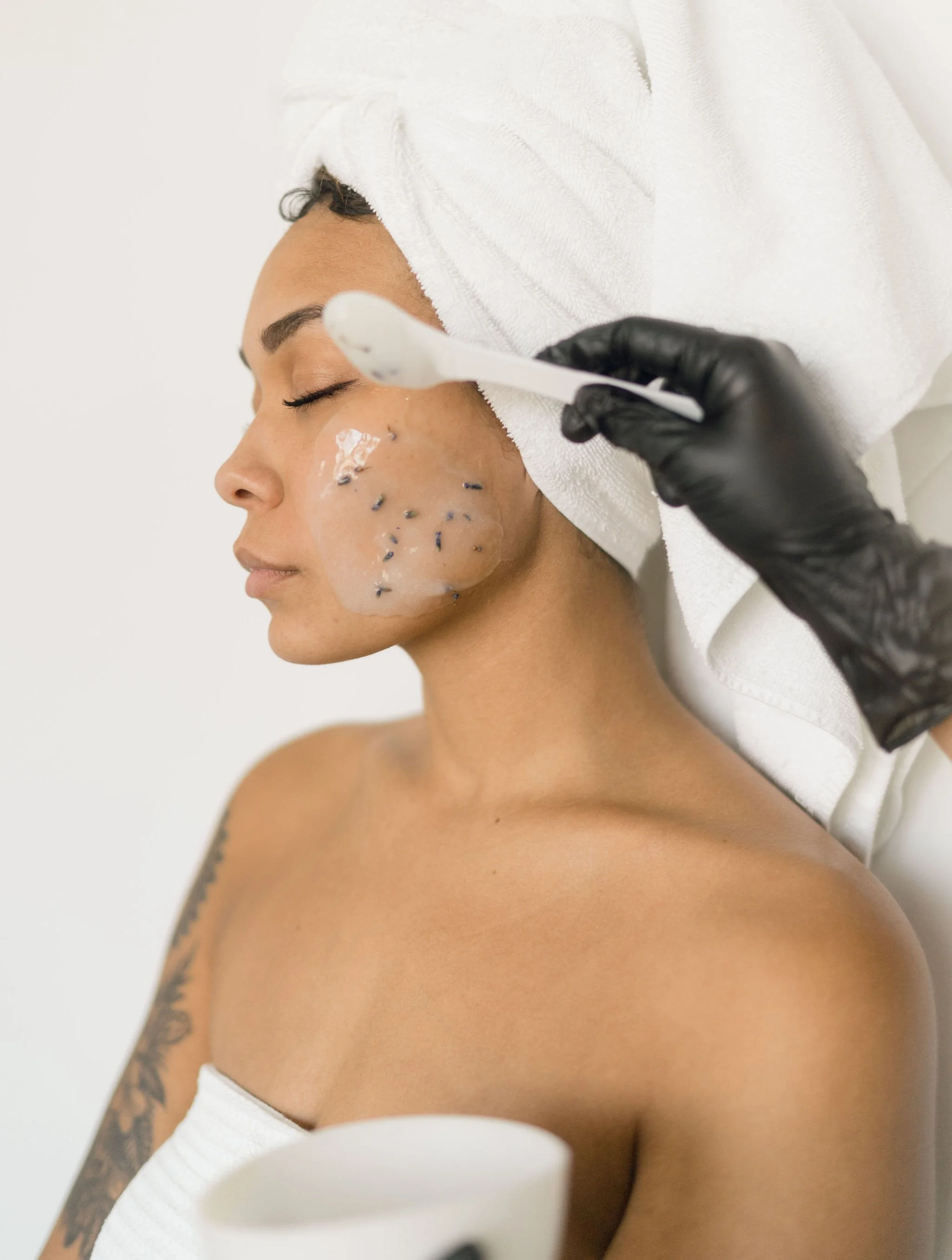 A woman receiving a facial treatment with a spatula tool, with small lavender buds on her cheek, covered in a facial mask, in a spa setting.
