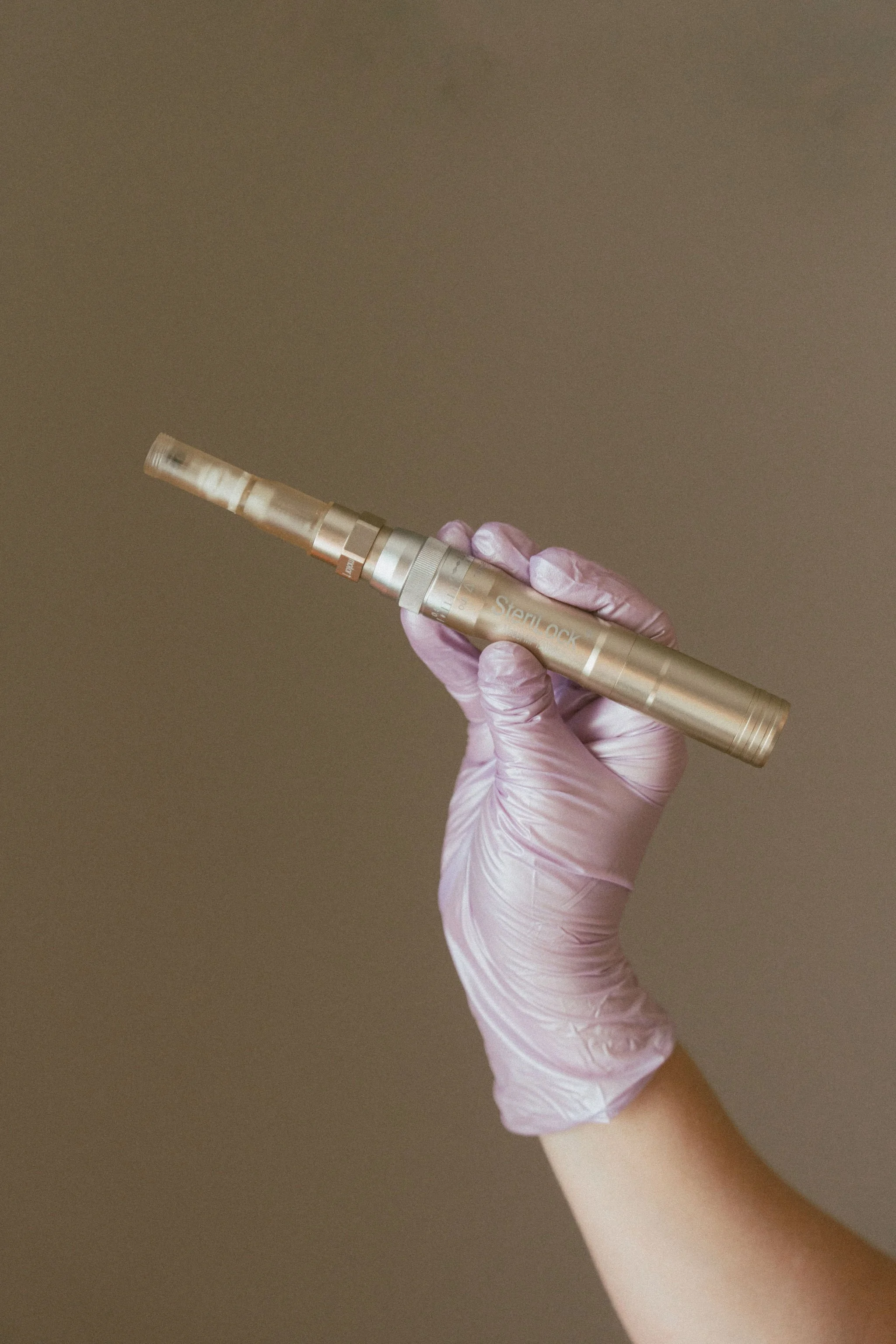 A hand wearing a pink medical glove holding a micro needling pen against a plain background.