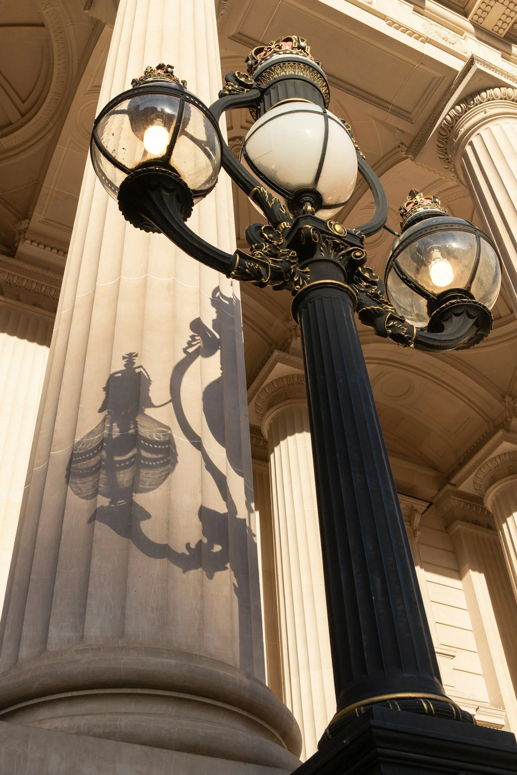 Close-up of an ornate black and gold street lamp with three glass globes, against a backdrop of classical architectural details including large columns and a decorated ceiling.