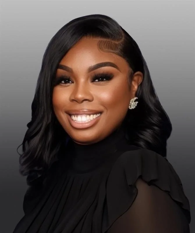 Smiling woman with black, wavy hair styled to the side, wearing a black top with ruffled shoulders, statement earrings, and makeup with bold eyebrows and lipstick.
