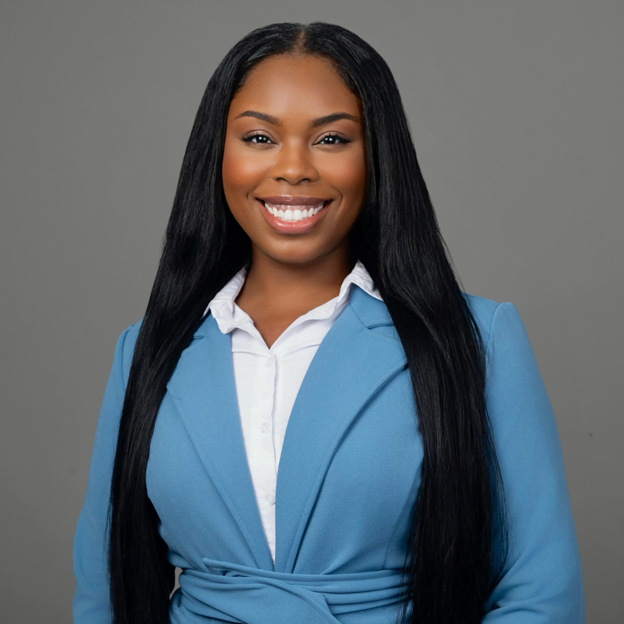 A young woman with long black hair, wearing a blue blazer and white shirt, smiling against a gray background.