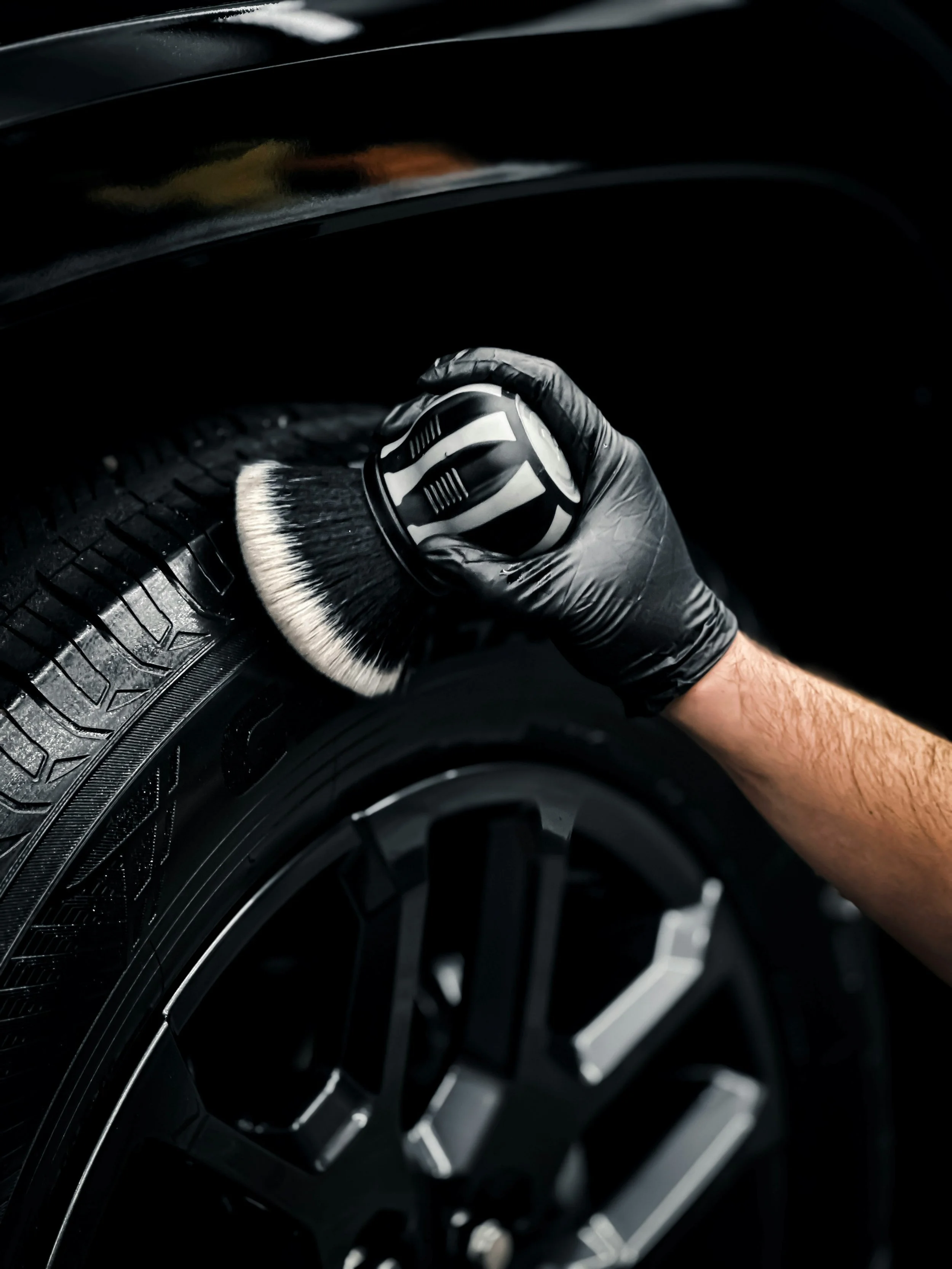 A person wearing a black glove is cleaning a black car tire with a brush that has a black and white handle.