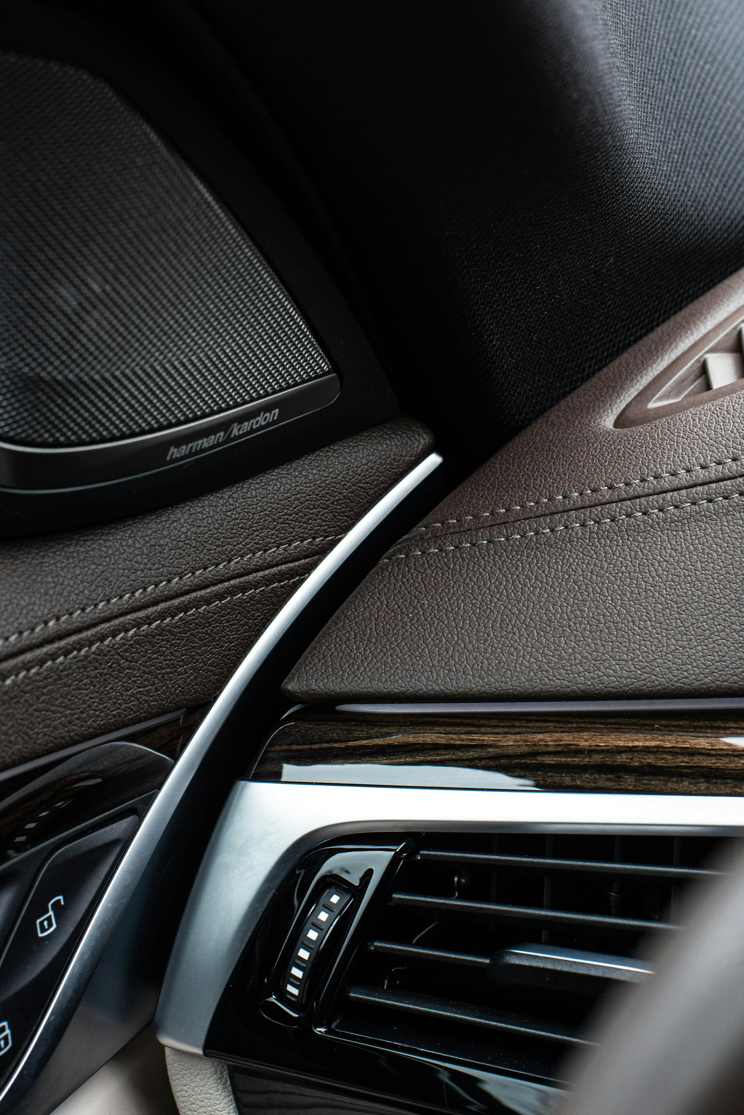 Close-up of the interior of a luxury car, showing a Harman Kardon speaker on the dashboard and part of the air conditioning vent, with brown leather upholstery and wood trim.