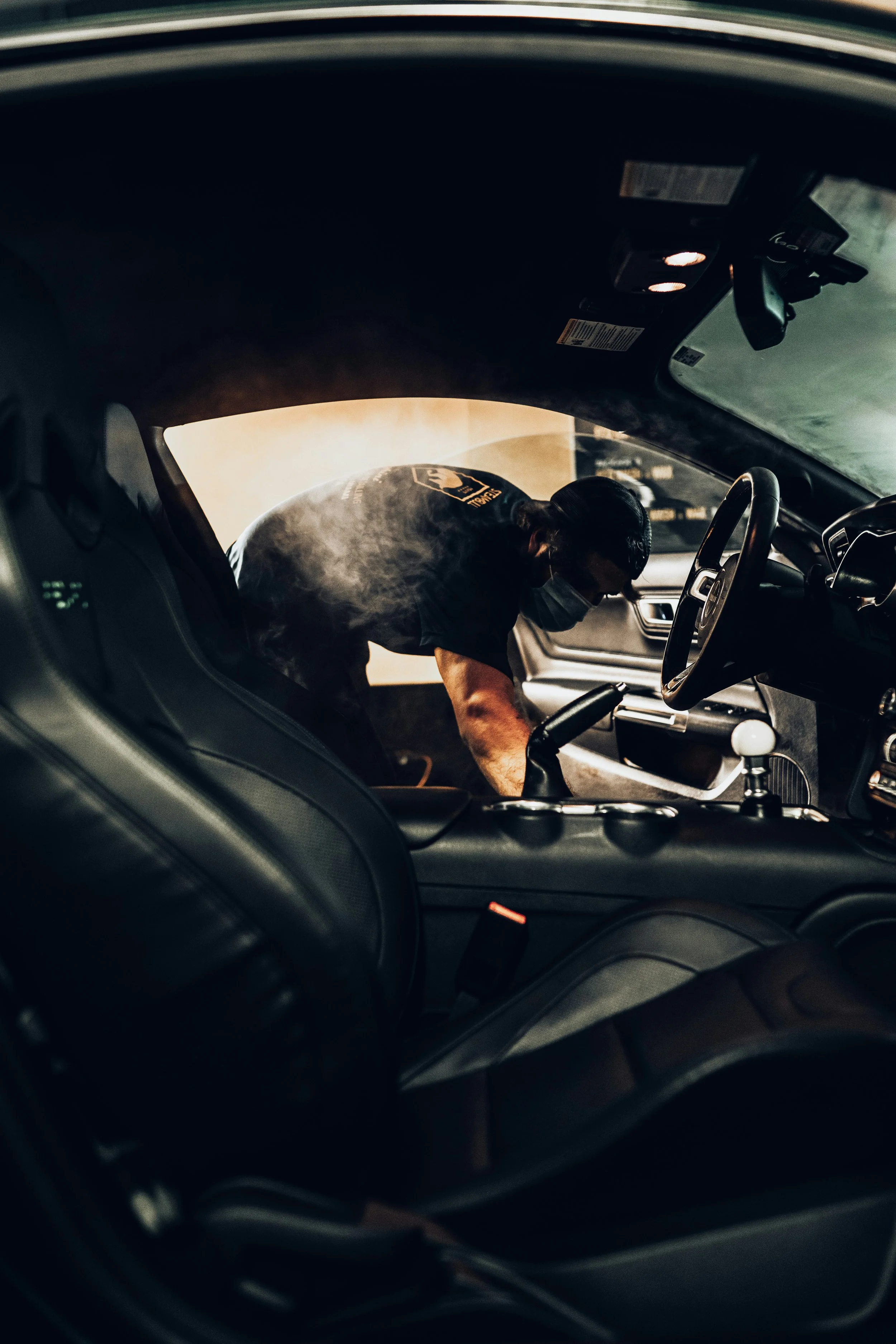 Person wearing a mask and cap, leaning into the passenger side of a car, inside a vehicle with a black interior.
