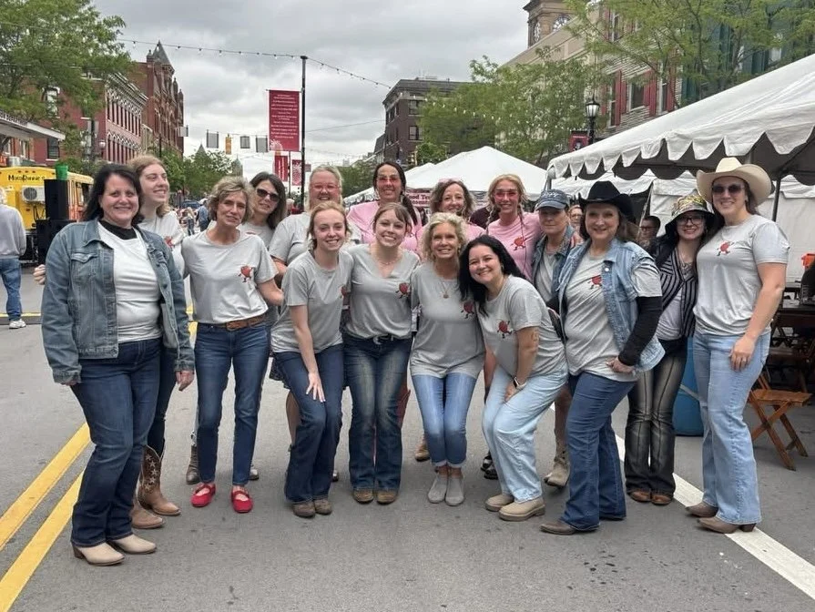 Group of women standing on a city street during a festival or outdoor event, with tents and buildings in the background, all wearing matching gray t-shirts with a red emblem.