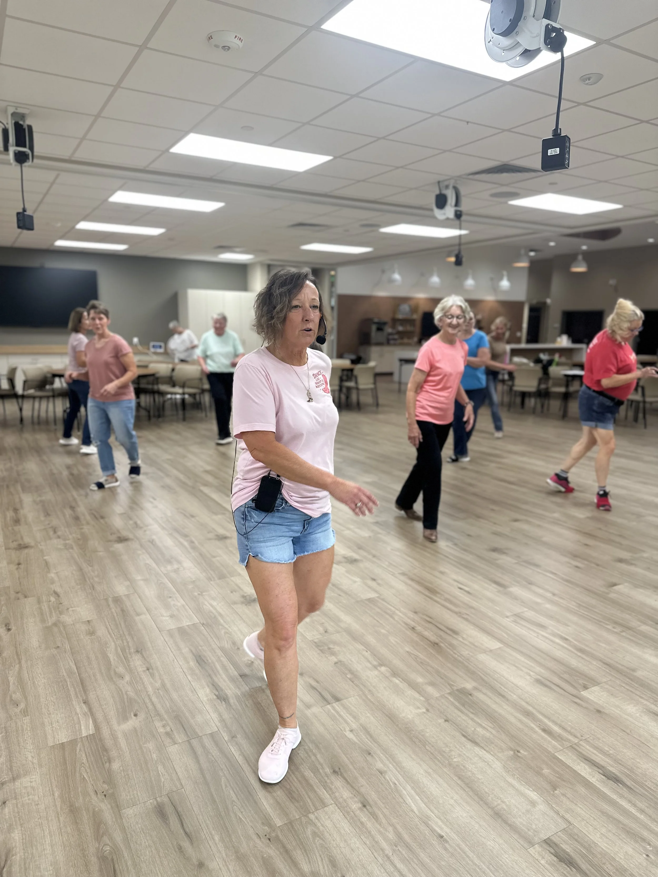 A group of women participating in a dance or exercise class in a studio with wooden floors and ceiling lights. One woman in the foreground is wearing a pink t-shirt, denim shorts, and white sneakers, and is wearing a headset microphone.