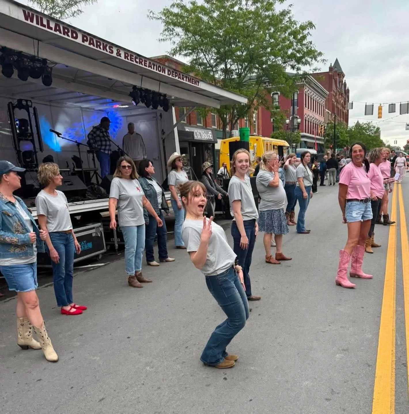A group of people dancing in front of a stage at an outdoor event, with a sign that reads 'Willard Parks & Recreation Department,' and a row of buildings on the street. The crowd is cheerful, and some people are wearing cowboy boots and pink shirts.