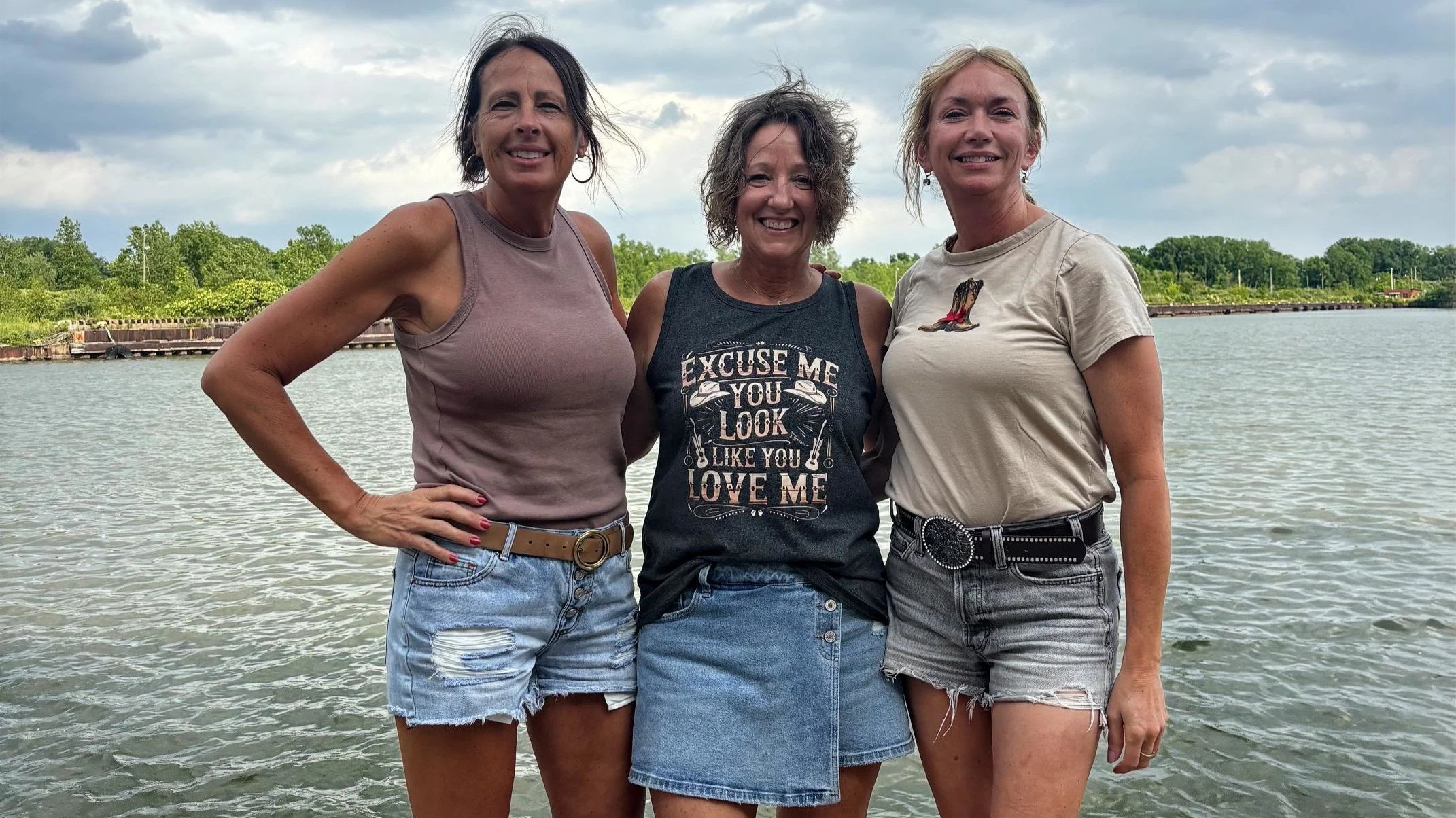 Three women standing close together outdoors near a body of water, smiling at the camera with trees and a cloudy sky in the background.